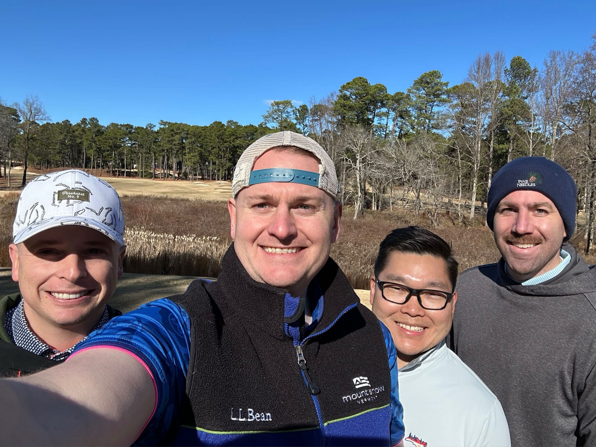Four people are smiling outdoors, standing in front of trees under a clear blue sky. They appear to be on a golf course.