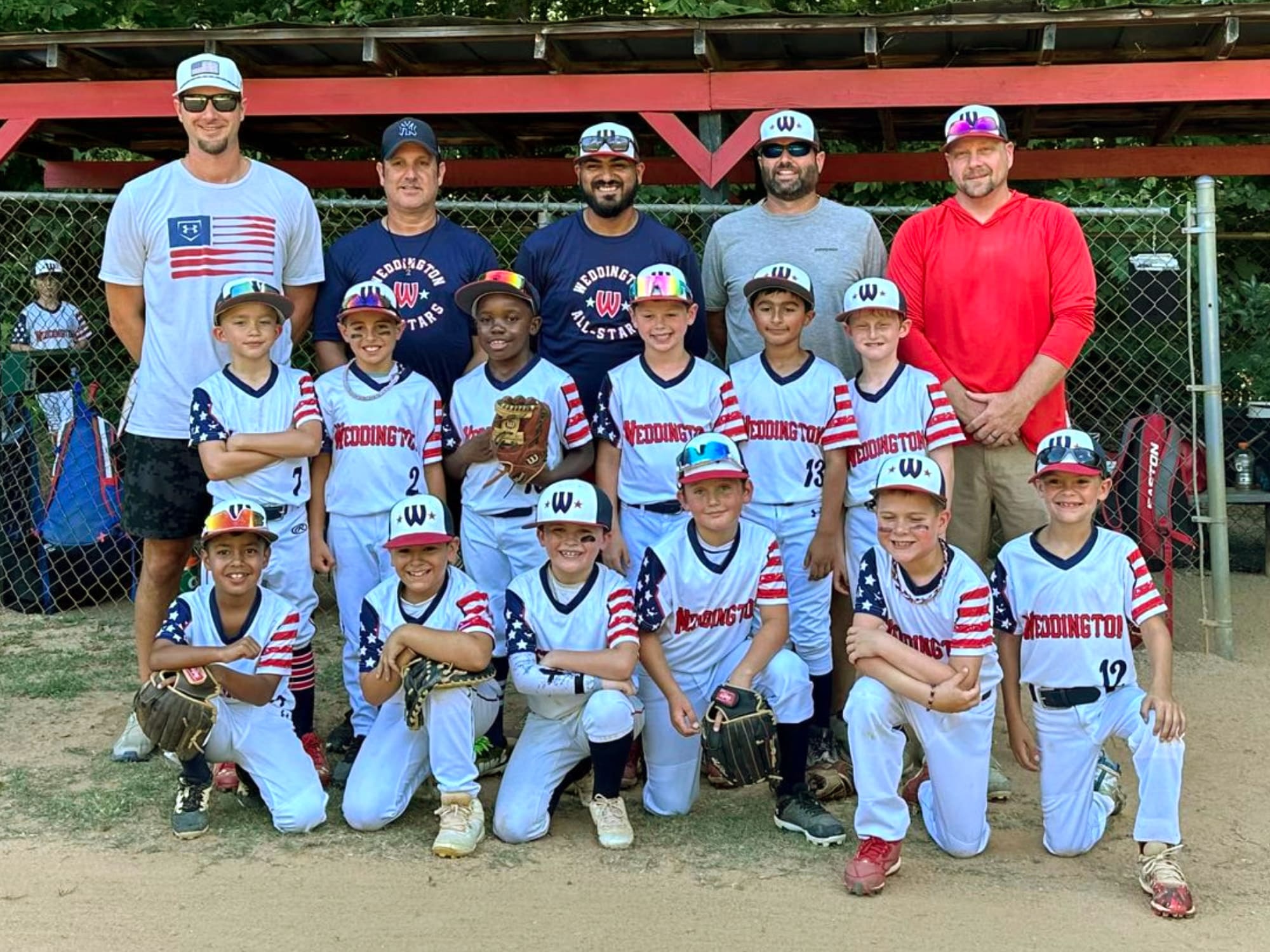 A youth baseball team poses together, featuring young players in Weddington uniforms and four adults, standing in front of a chain-link fence.