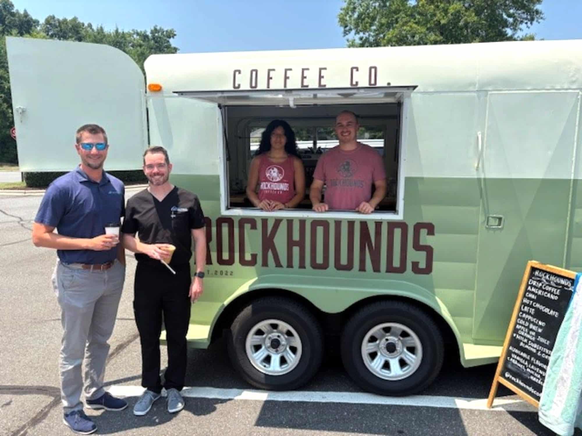 Three people with a coffee truck labeled "Rockhounds." Two individuals stand outside, two inside. Menu board displays coffee options. Sunny day with greenery.