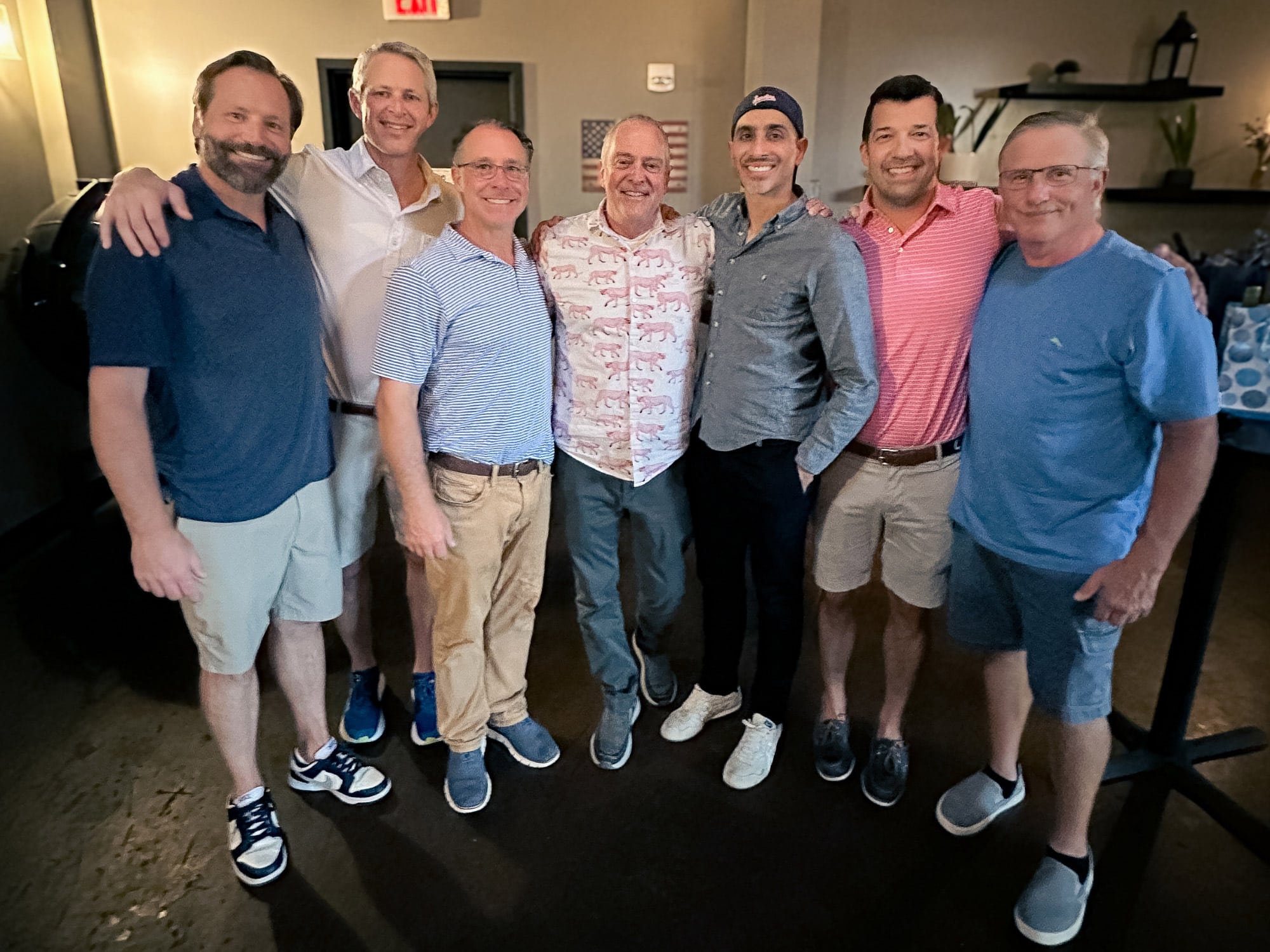 Seven people smiling and posing together indoors, casual attire, relaxed atmosphere. Dim lighting with shelves and decor in the background, suggesting a social gathering.