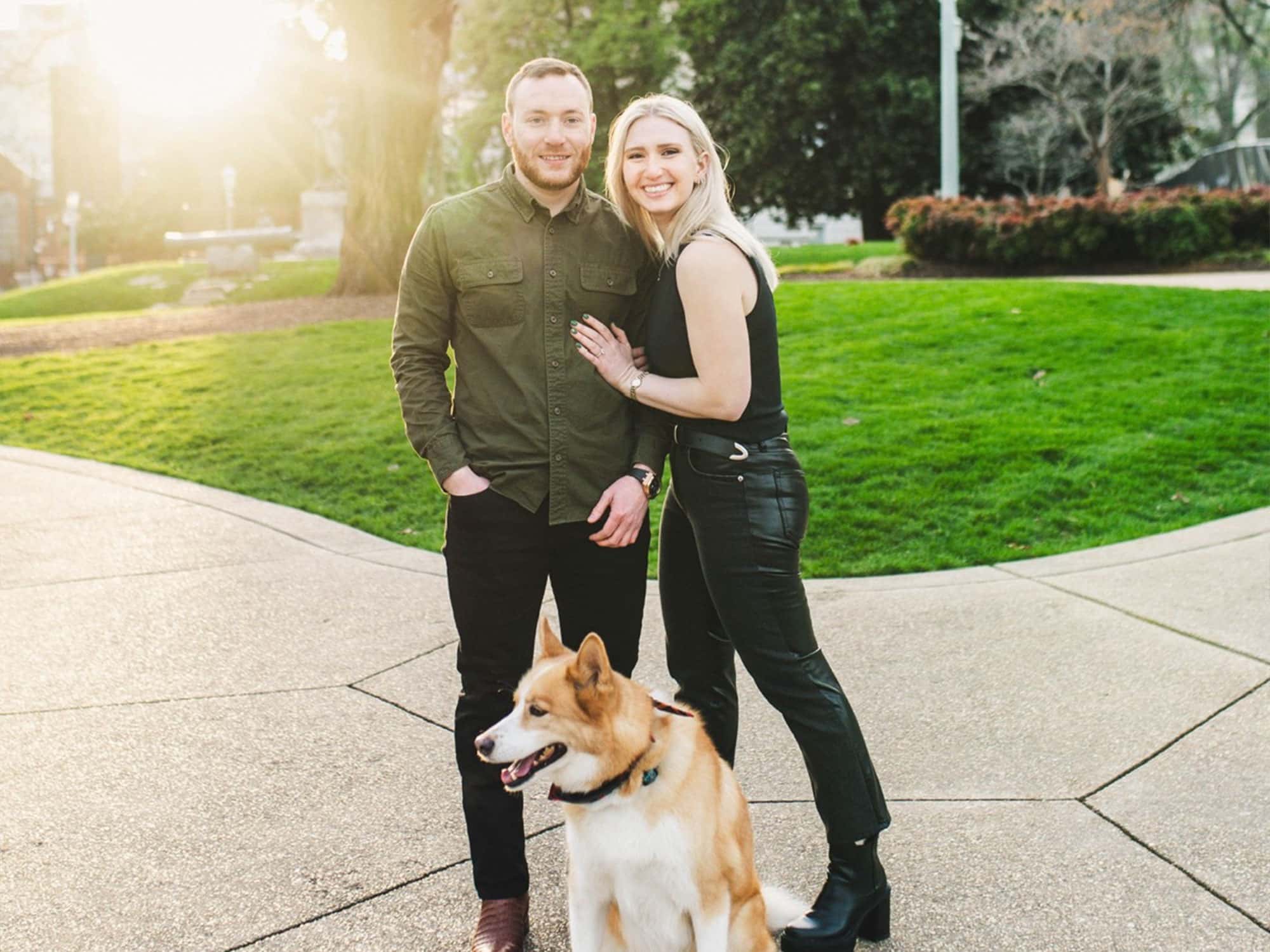 A person couple poses with a dog on a sunny day in a park, surrounded by greenery and a paved pathway.