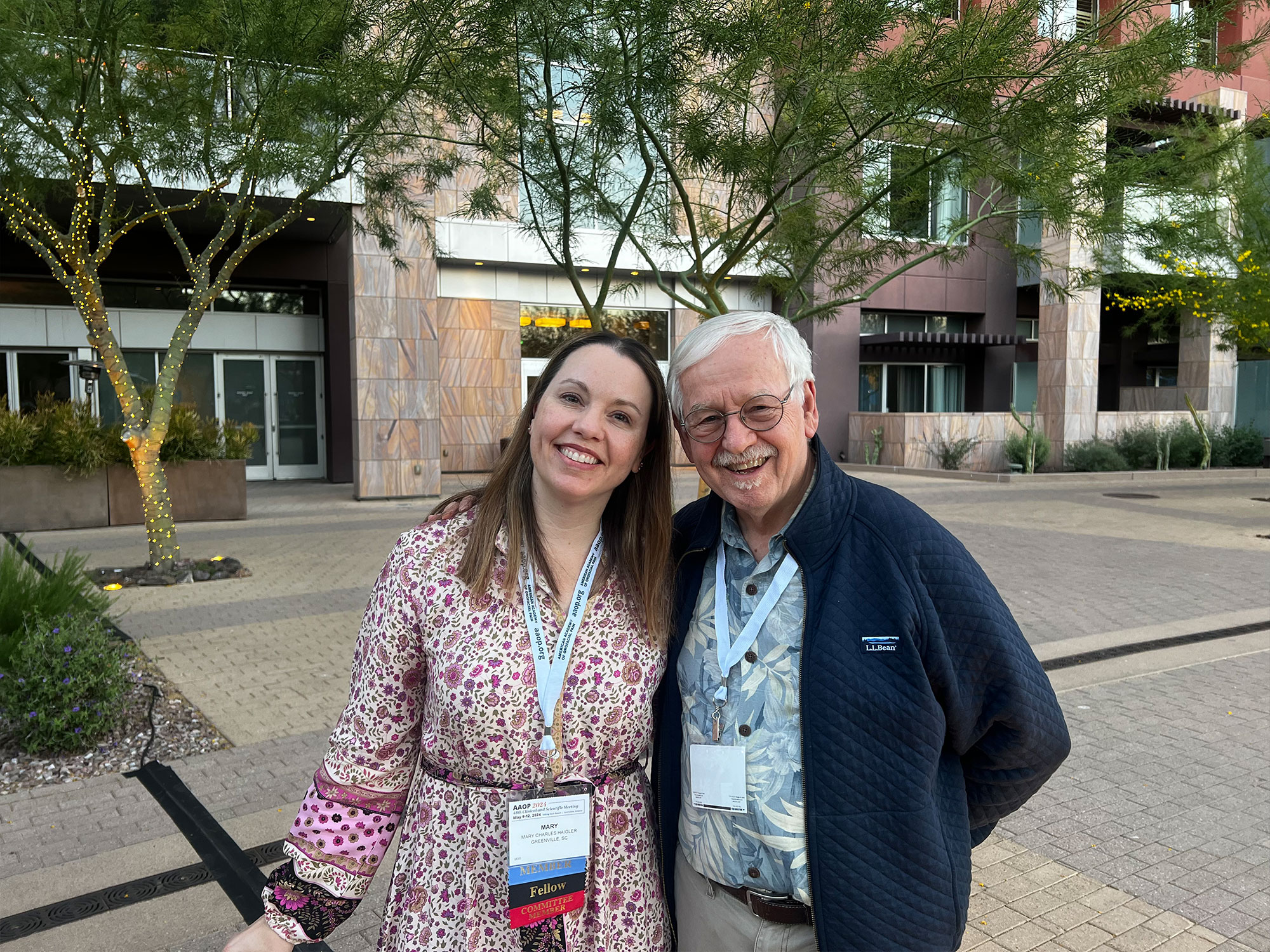Two people smile outdoors near a modern building entrance with decorative trees. Both wear conference badges, suggesting a professional event or gathering.