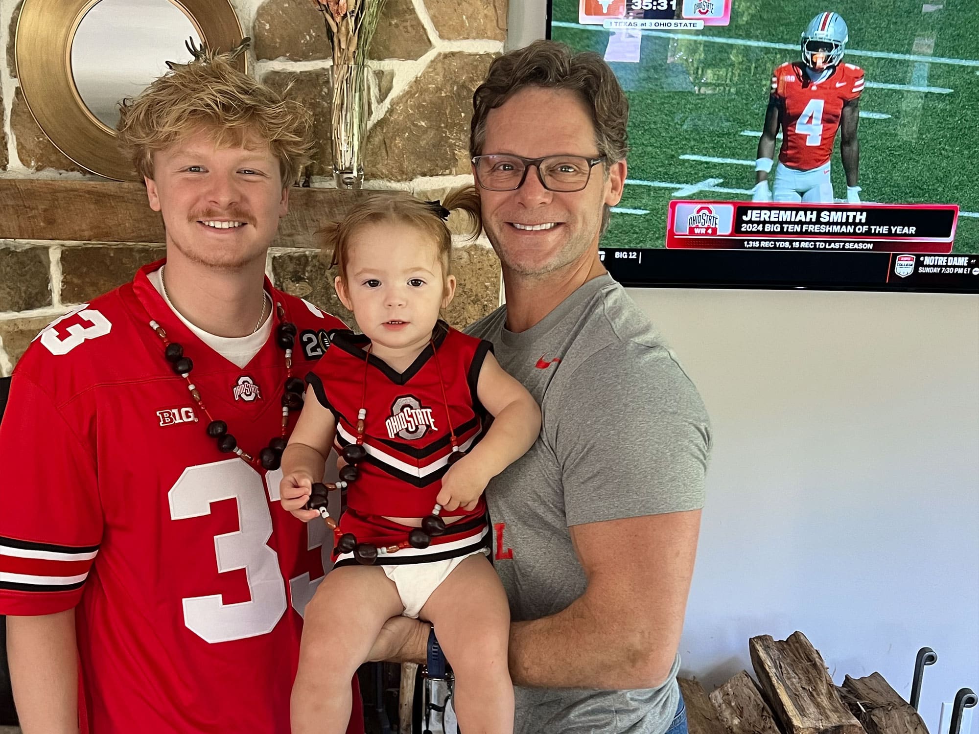 A person holds a child near a stone fireplace. Another person stands nearby wearing an Ohio State jersey, with a football game on TV.