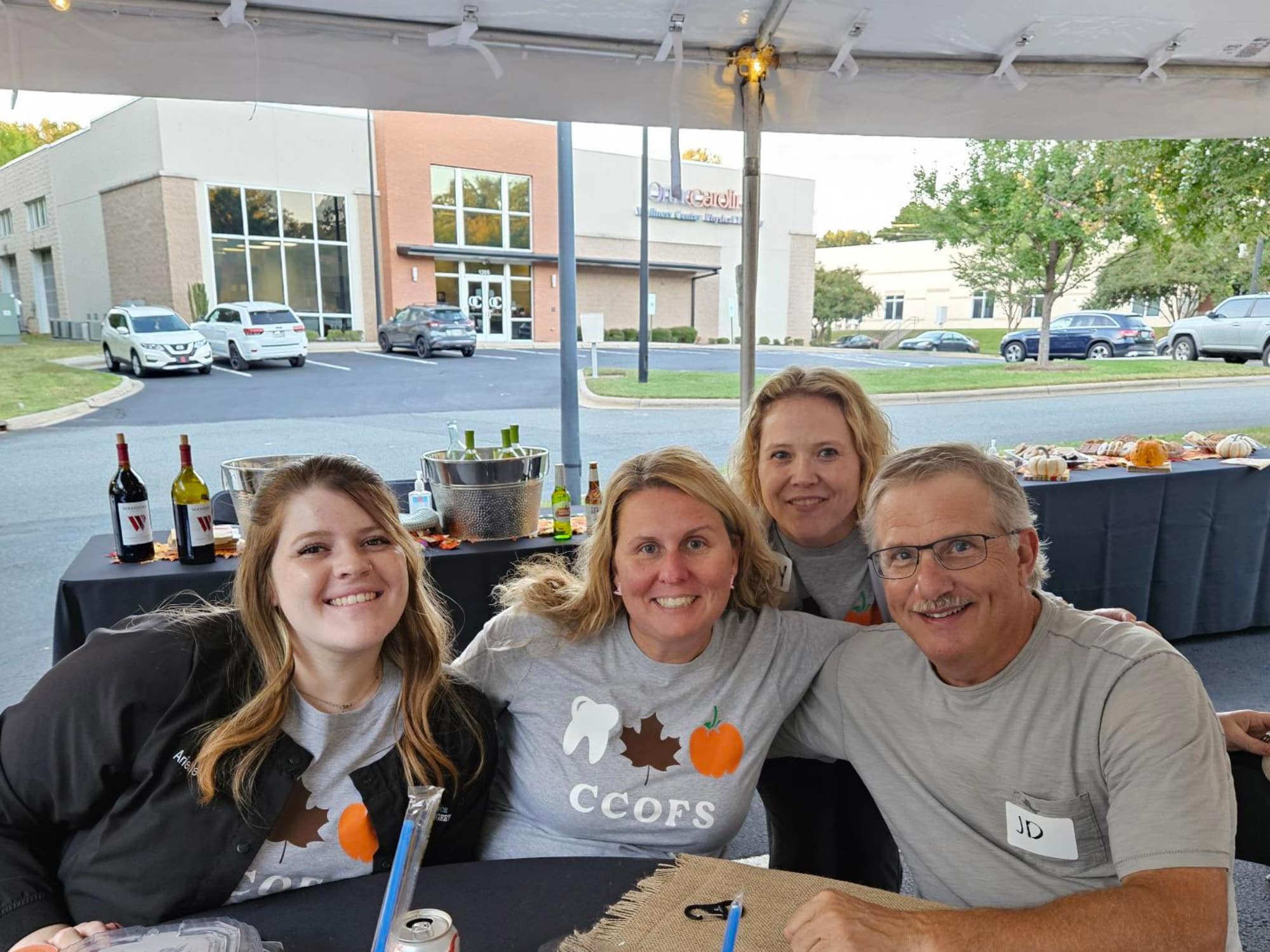 Four people smiling under a tent with a table of beverages and snacks. Buildings and parked cars are visible in the background.