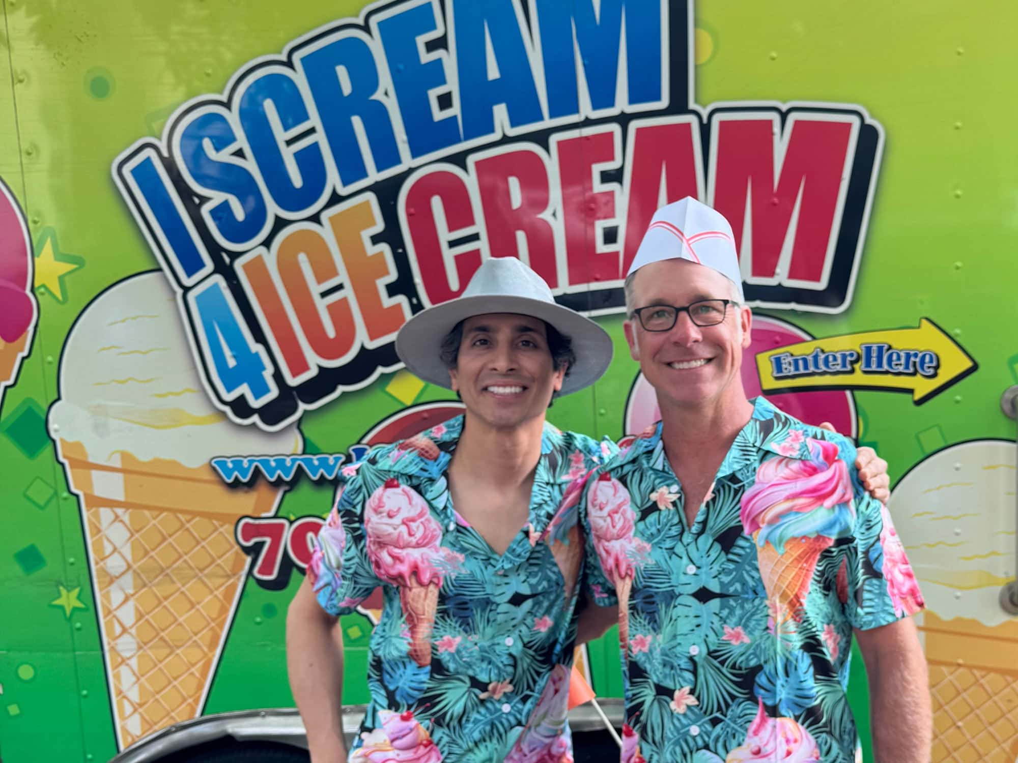 Two people in colorful shirts stand smiling in front of an ice cream truck with vibrant signage.