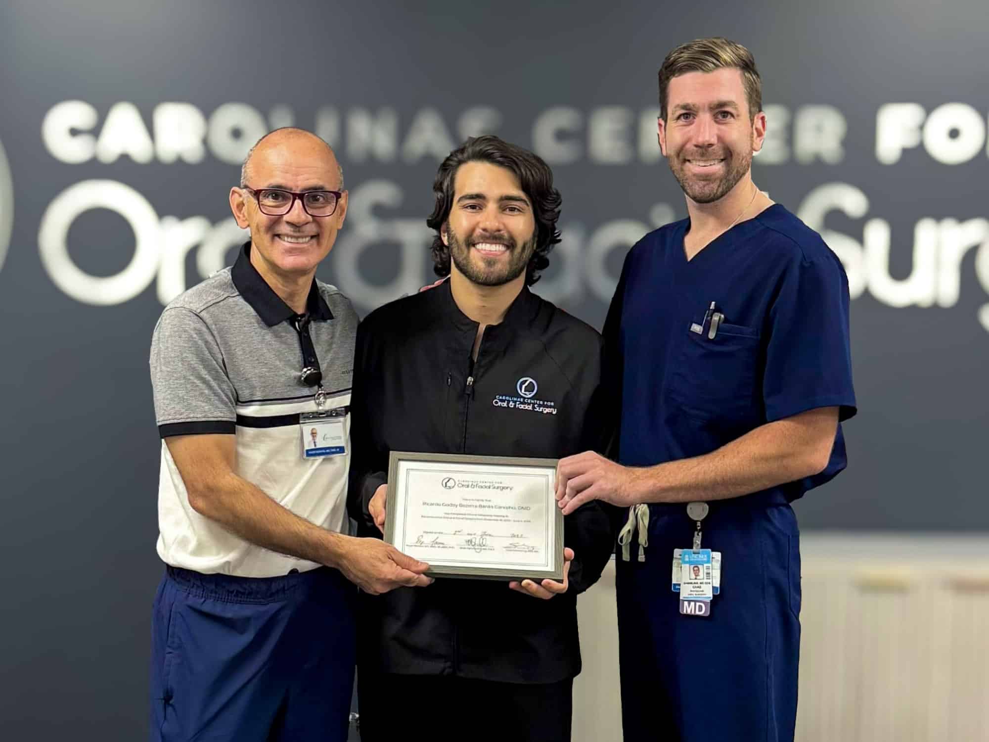 Three people smiling, holding a certificate in front of a sign for Carolinas Center for Oral and Facial Surgery. Casual and professional attire.