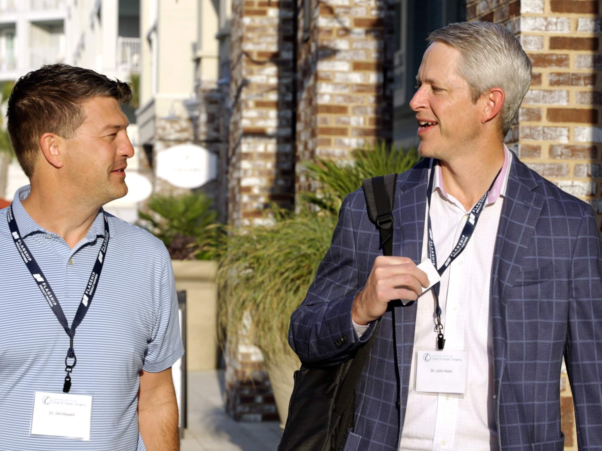 Two people with name badges walk outdoors, engaging in conversation. The setting features brick buildings and ornamental plants, suggesting a professional event.