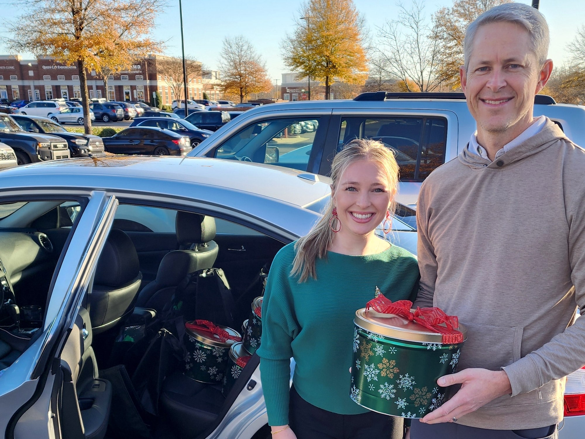 Two people smiling near a car outside. They're holding a festive tin. Background includes parked cars and a brick building with "Queen City" sign.