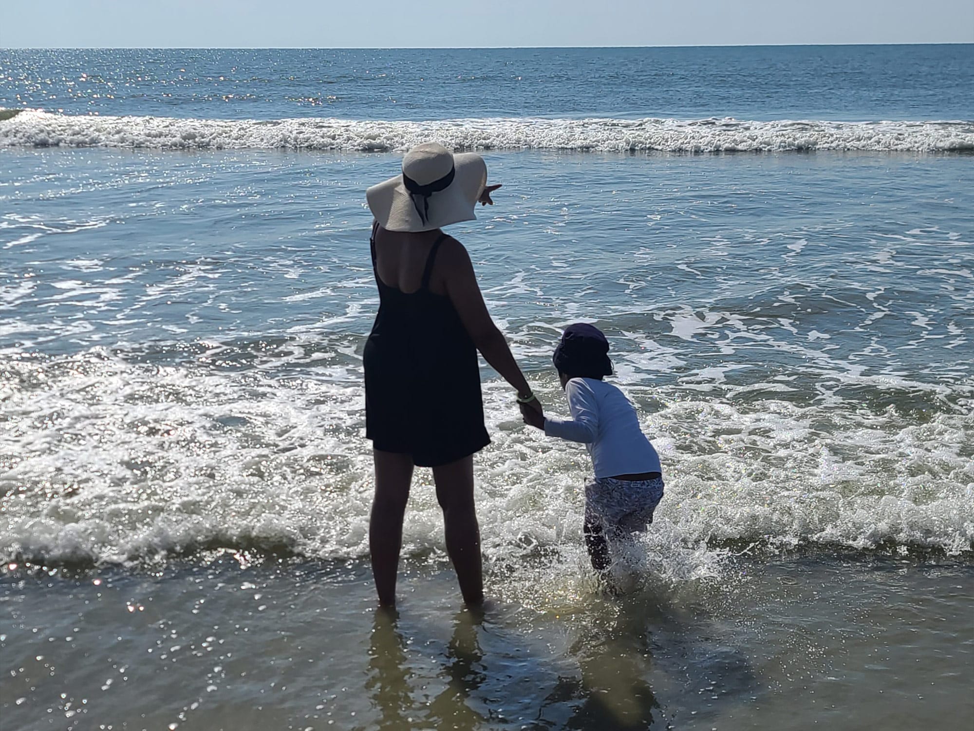 A person and child stand in the ocean waves, facing the horizon, as the person points towards the distance on a sunny day.