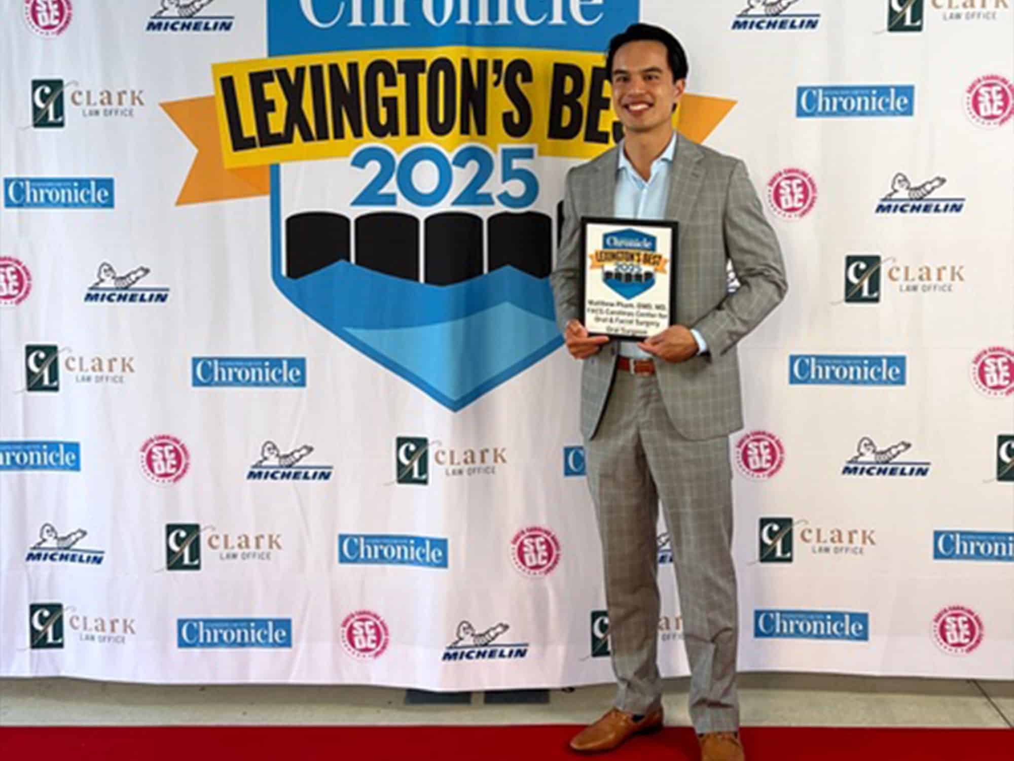 A person in a grey suit holds a certificate in front of a "Lexington's Best 2025" banner, with various sponsor logos.