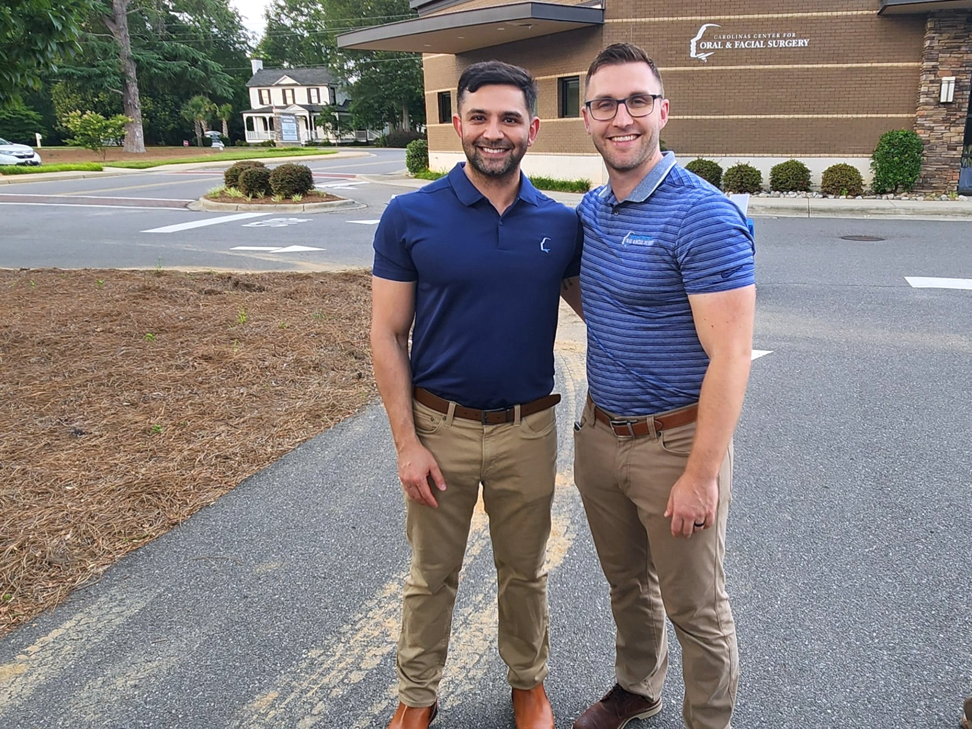 Two people smiling, standing outdoors near a medical facility sign, "Carolinas Center for Oral & Facial Surgery," with trees and a house in the background.