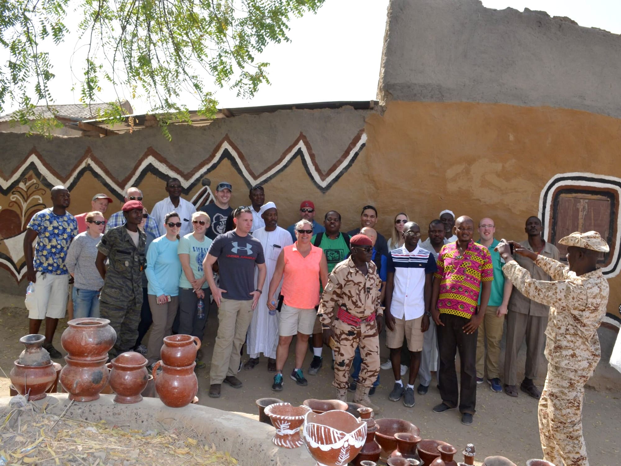 A group of people poses in front of a traditionally painted mud house. Earthen pots are displayed in the foreground.
