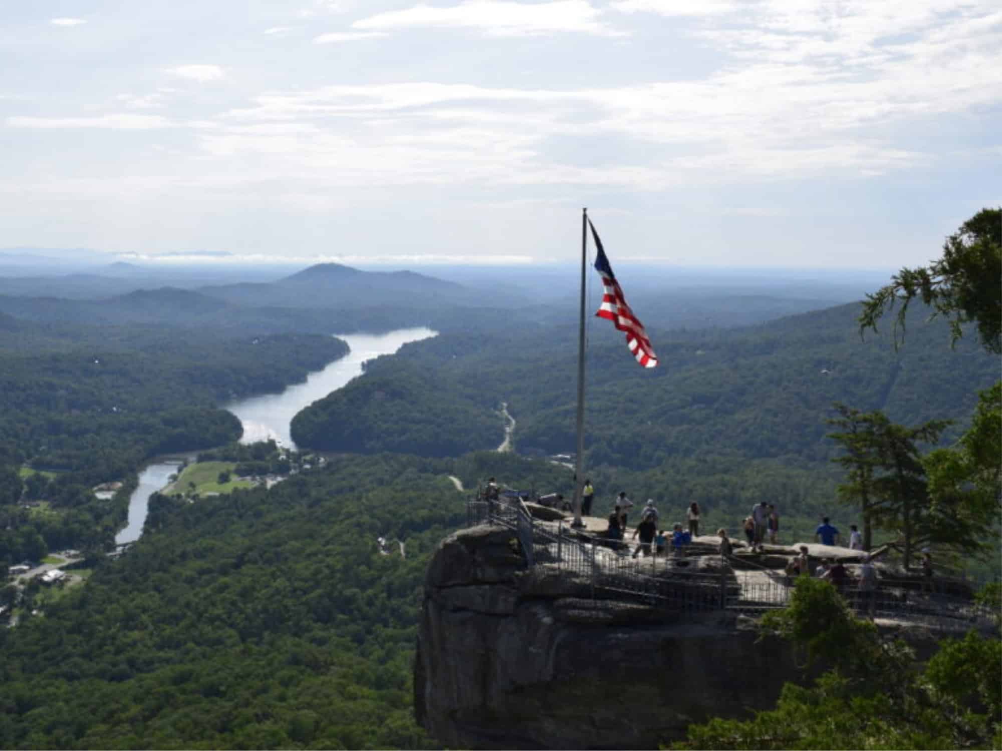 A group of people at Chimney Rock with an American flag, overlooking a lush valley and winding river under a vast sky.
