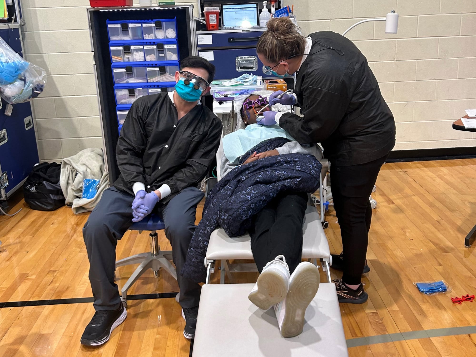 Two people in medical attire assist a patient on a folding chair. Supplies are organized in a portable cabinet. Indoor setting.