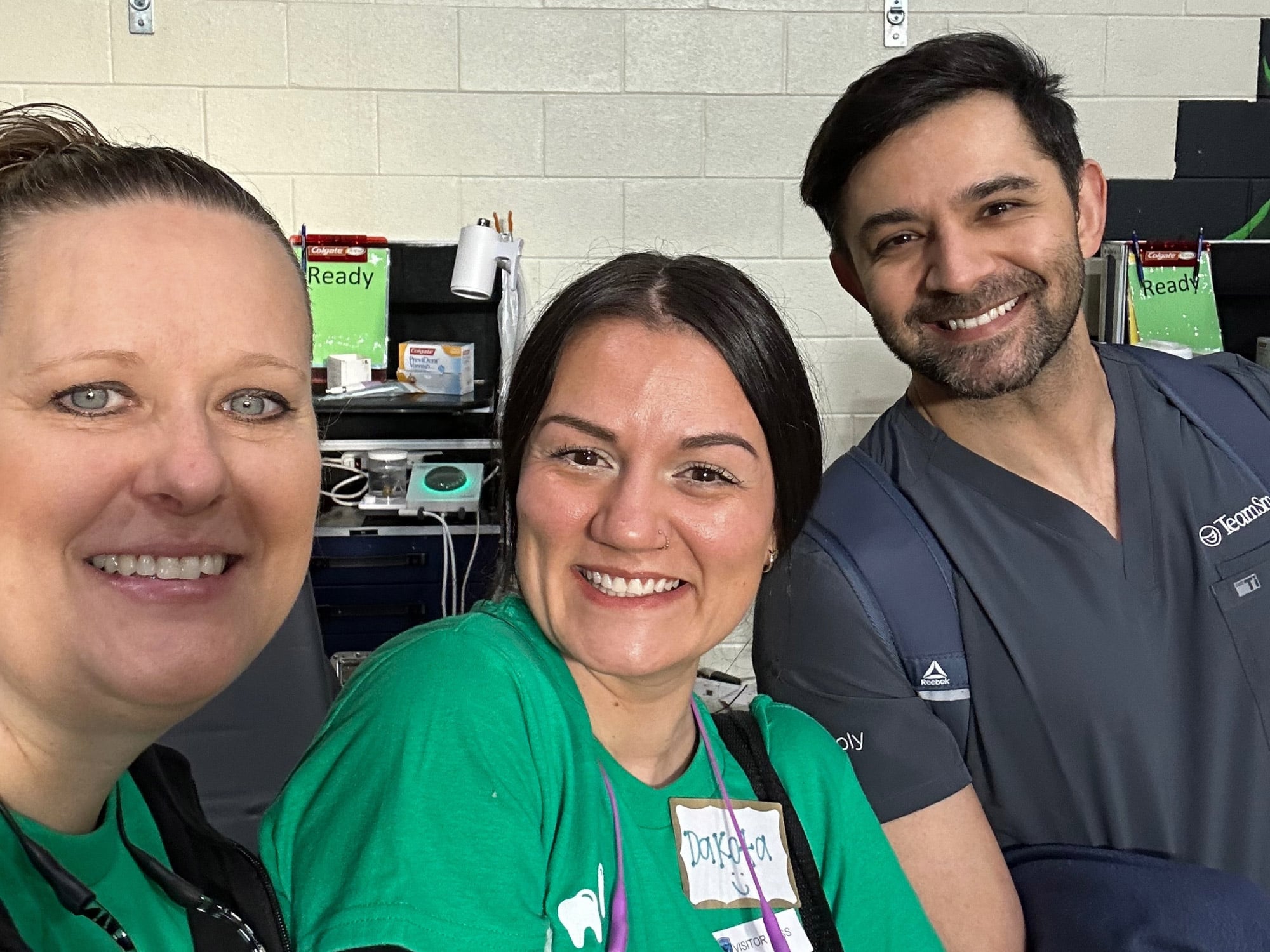 Three people smiling indoors, wearing casual and professional attire. Background features equipment and supplies, suggesting a healthcare or dental setting.