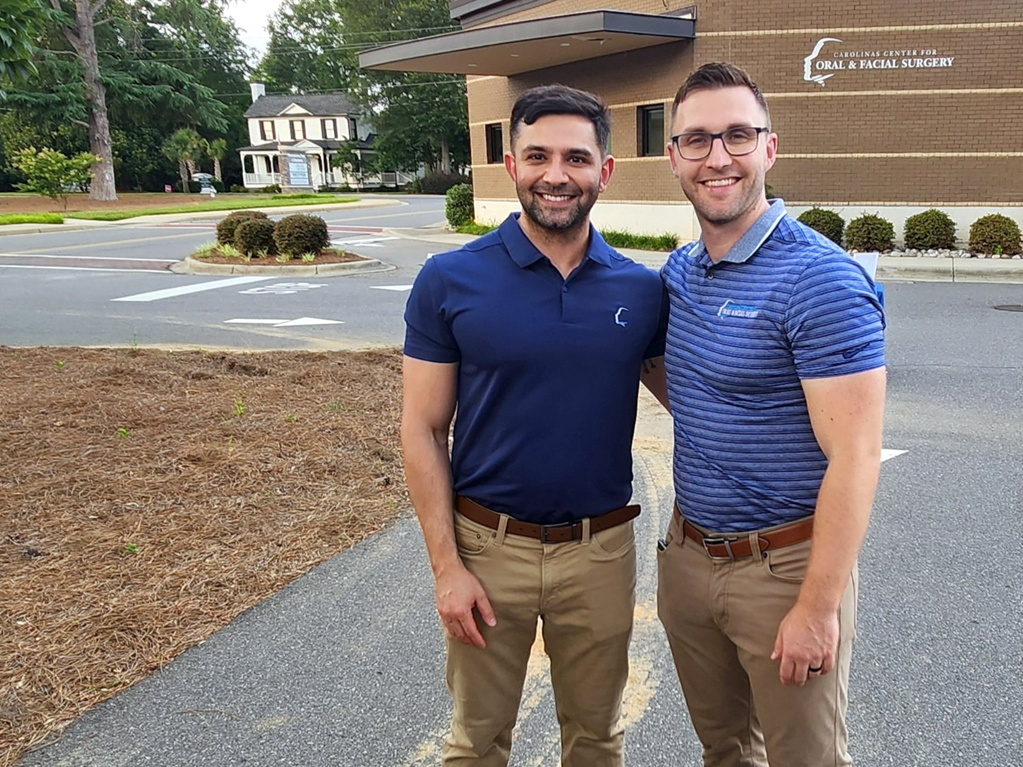 Two smiling persons standing outside Carolinas Center for Oral & Facial Surgery, with trees and a brick building in the background.