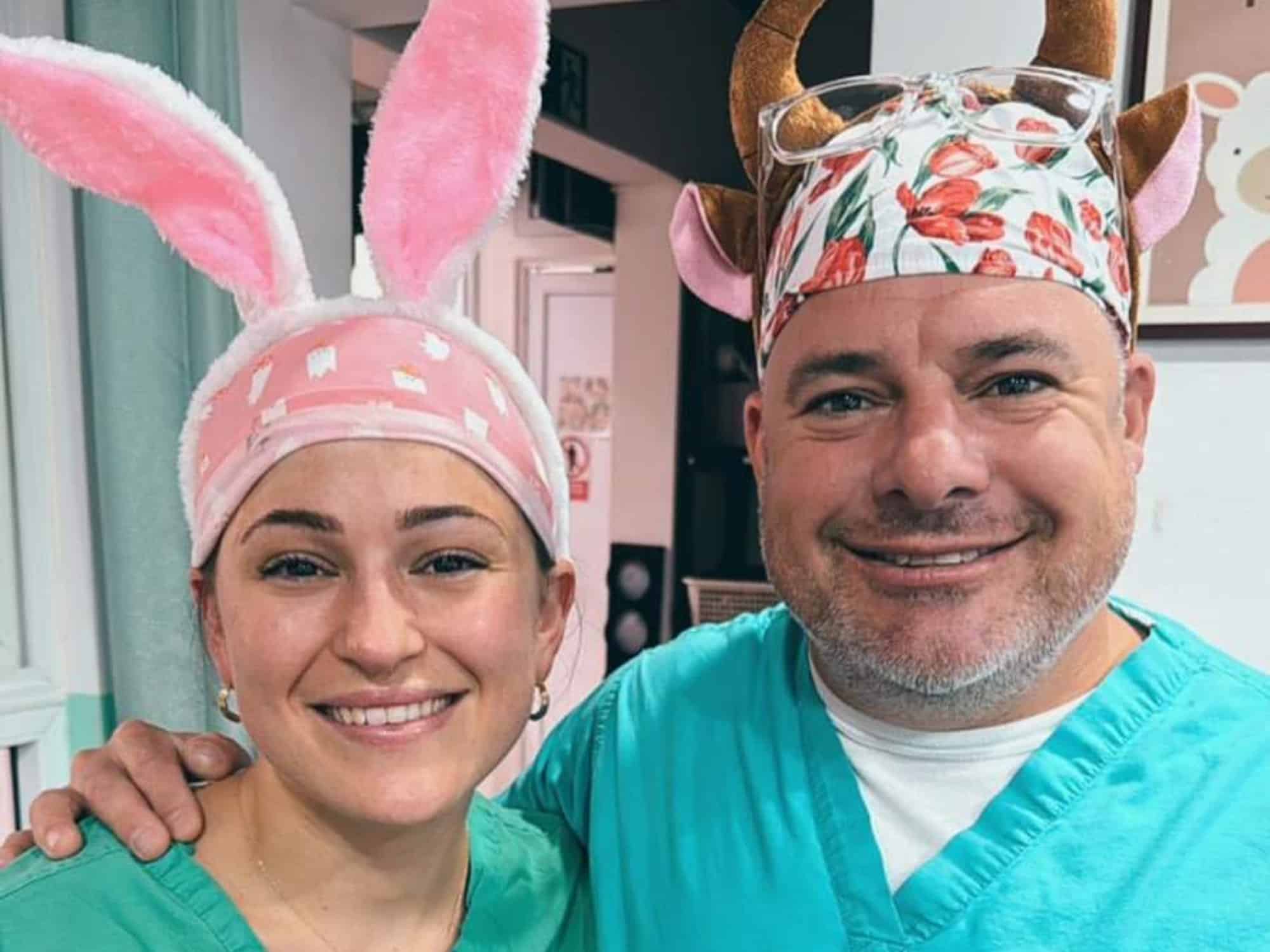 Two people in scrubs, smiling, wearing whimsical headbands with bunny ears and floral horns, in a cheerful room with colorful decor.
