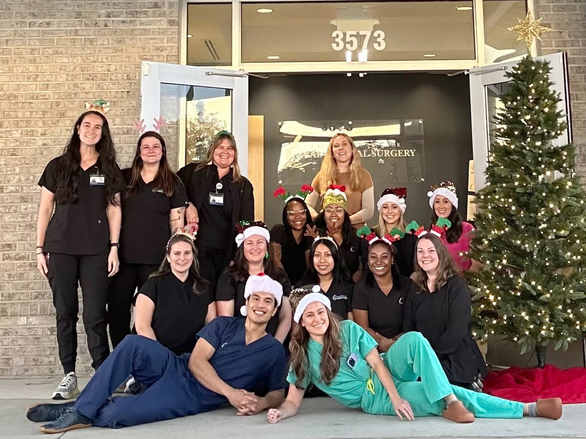 A group of people in festive attire pose outside a building numbered 3573, next to a decorated Christmas tree.