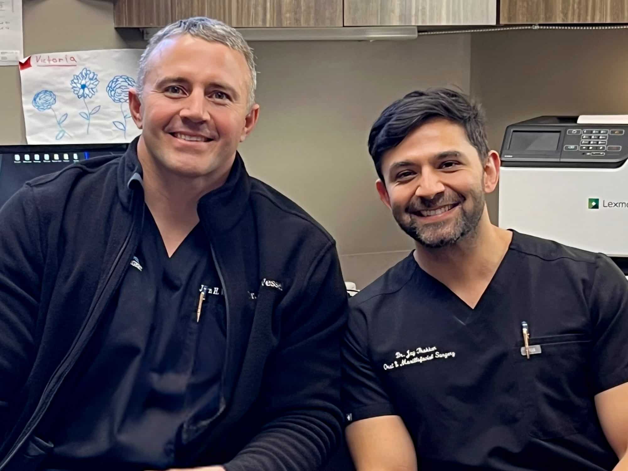 Two people smiling in a professional setting, wearing black outfits with embroidered names. A child’s drawing is displayed on the wall.