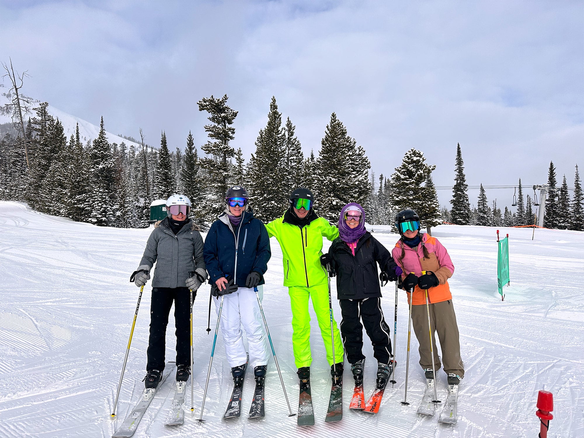 Five people in ski gear pose on a snowy slope, surrounded by evergreen trees and mountains on a cloudy day.