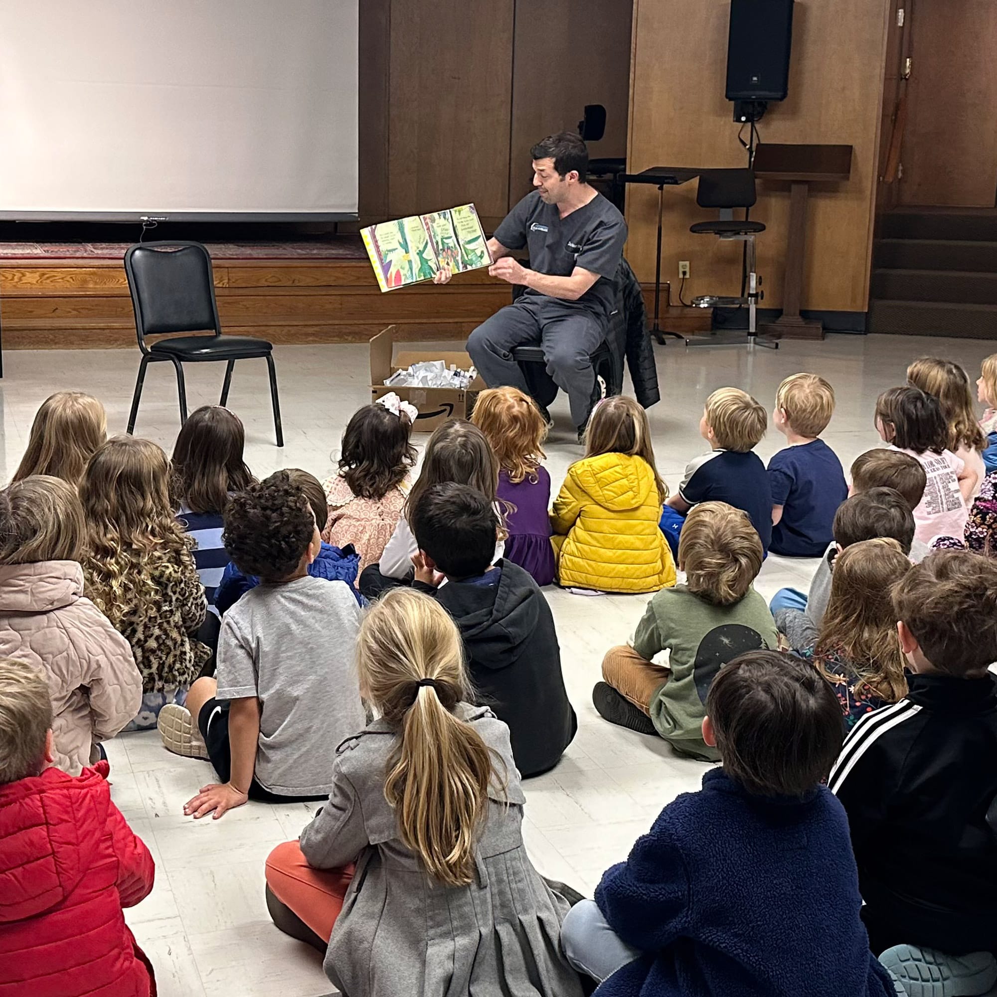 A person reads a story to a group of attentive children seated on a classroom floor, with a chair nearby.