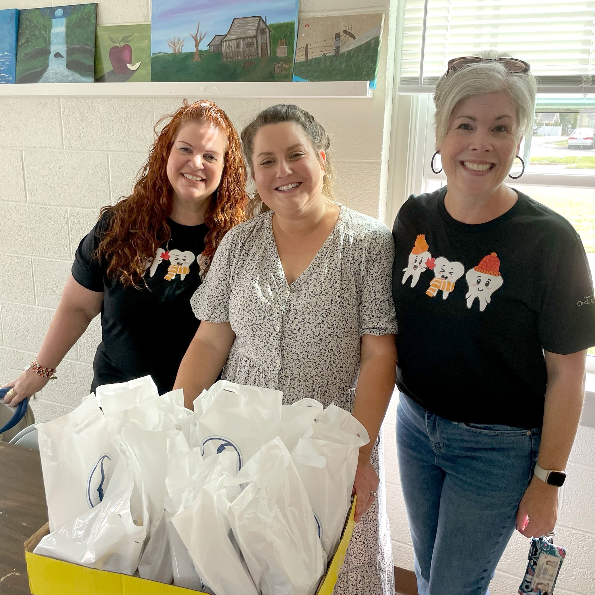 Three people smiling indoors with a box of white bags. Paintings decorate the wall behind them, and sunlight enters through the window.