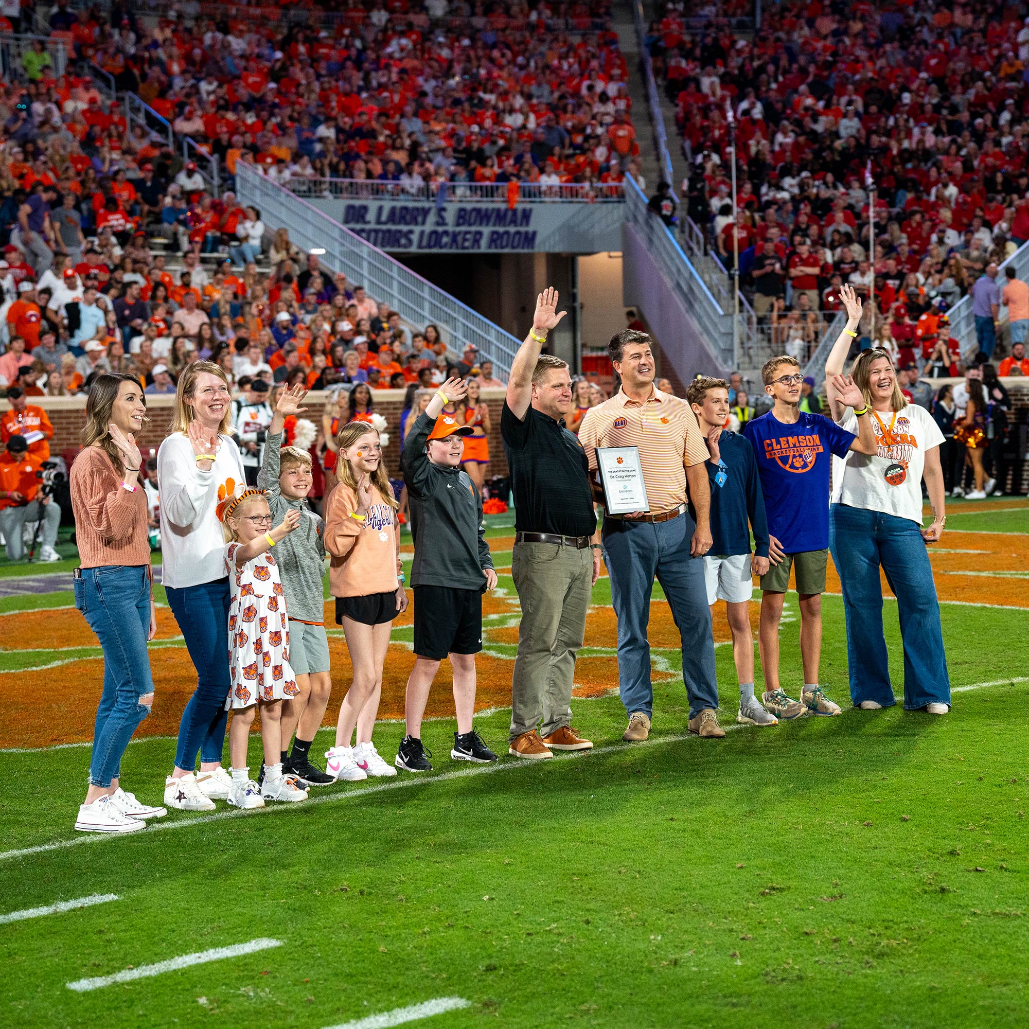 A group on a football field waves to a stadium audience. Some individuals hold a framed certificate. The environment is lively and colorful.