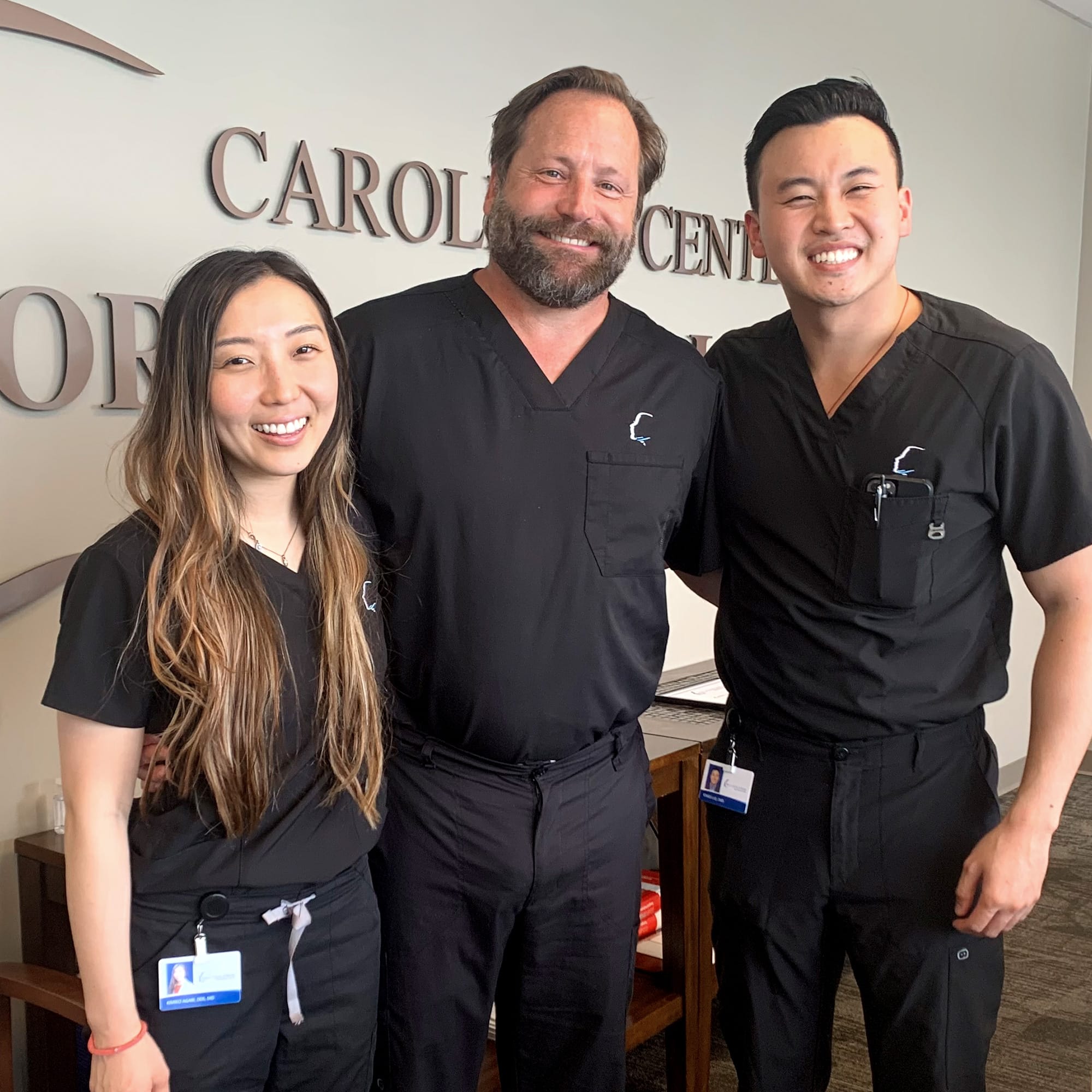 Three people in black medical scrubs smile in an office setting, next to a wall with partial text "CAROLI...".