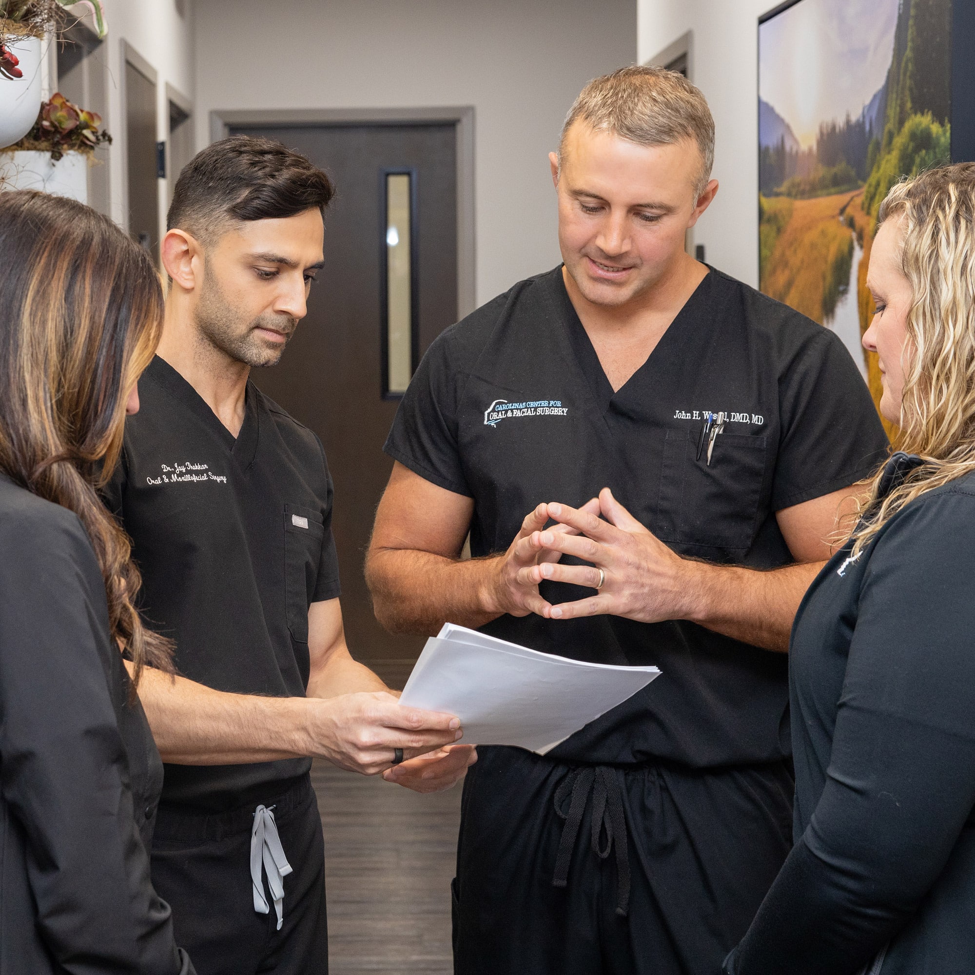 Four people in black scrubs are discussing papers in a hallway. They're likely in a medical or professional setting.