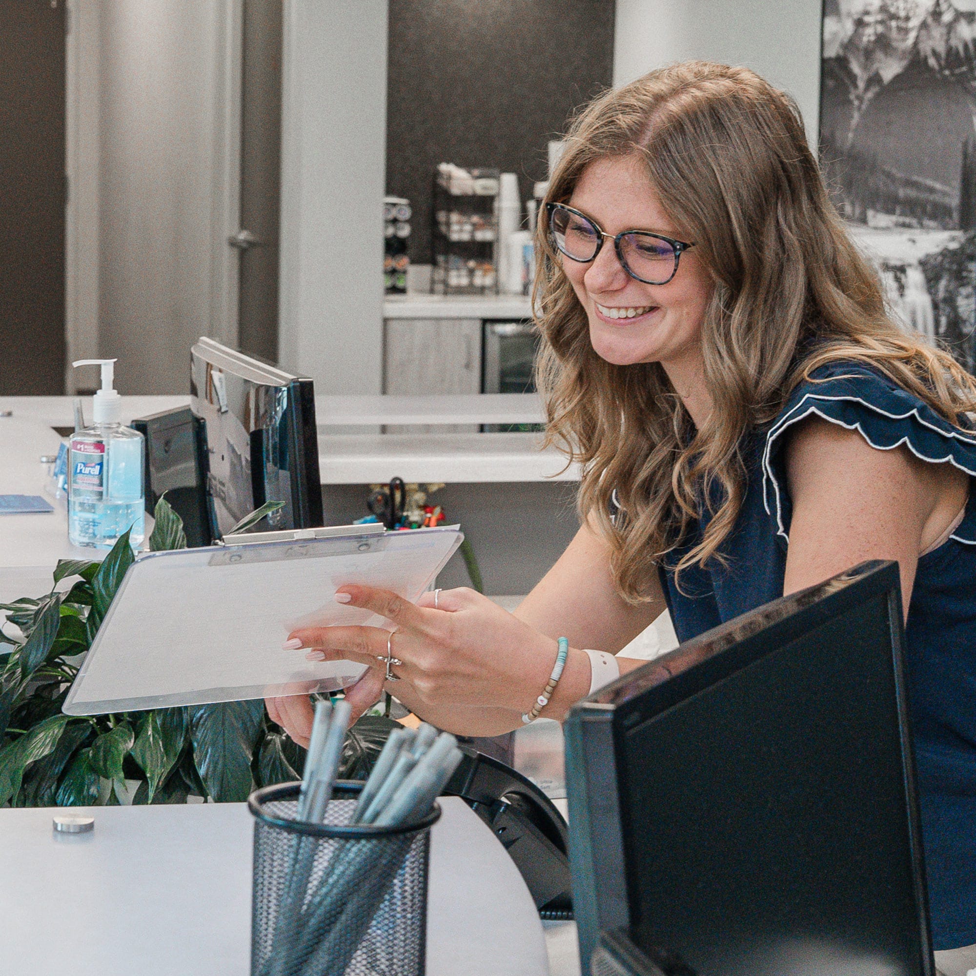 A person smiles while holding a clipboard in an office setting. Nearby are desks, plants, and office supplies, including pens and hand sanitizer.