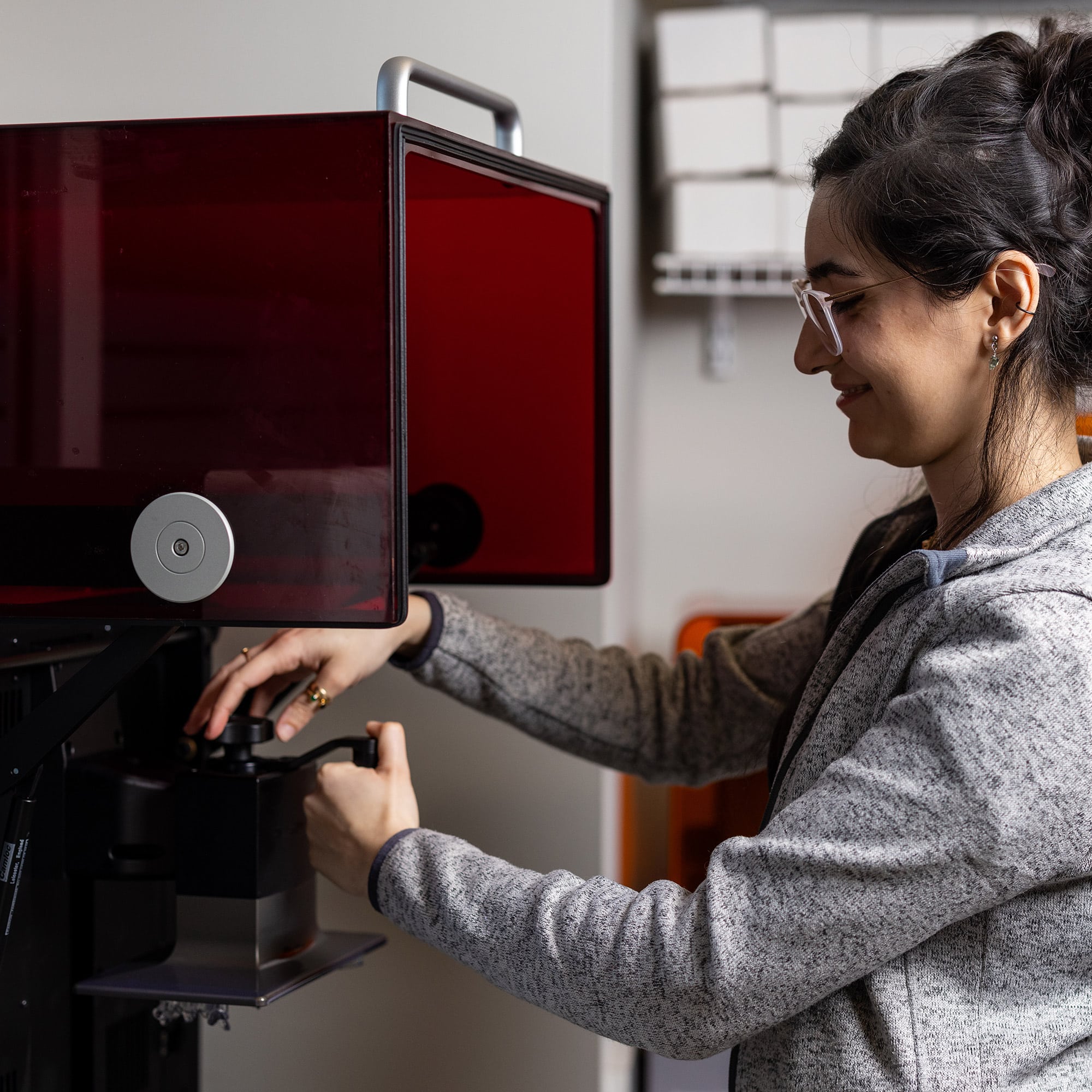 A person operates a 3D printer in a workshop, focusing intently. Shelves with organized boxes are visible in the background.
