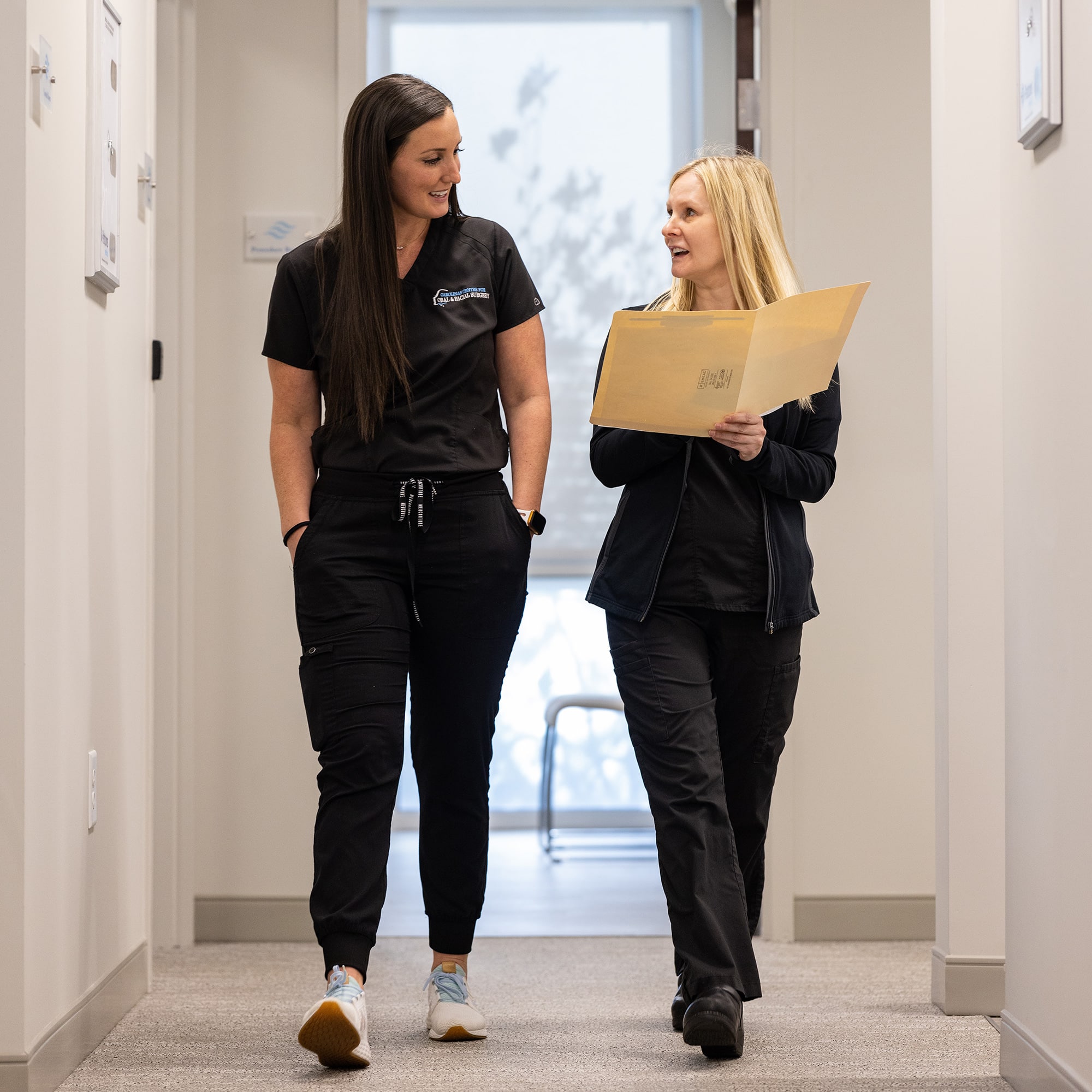 Two persons in black scrubs walk down a hallway, discussing documents in a professional setting. Both appear engaged and focused.