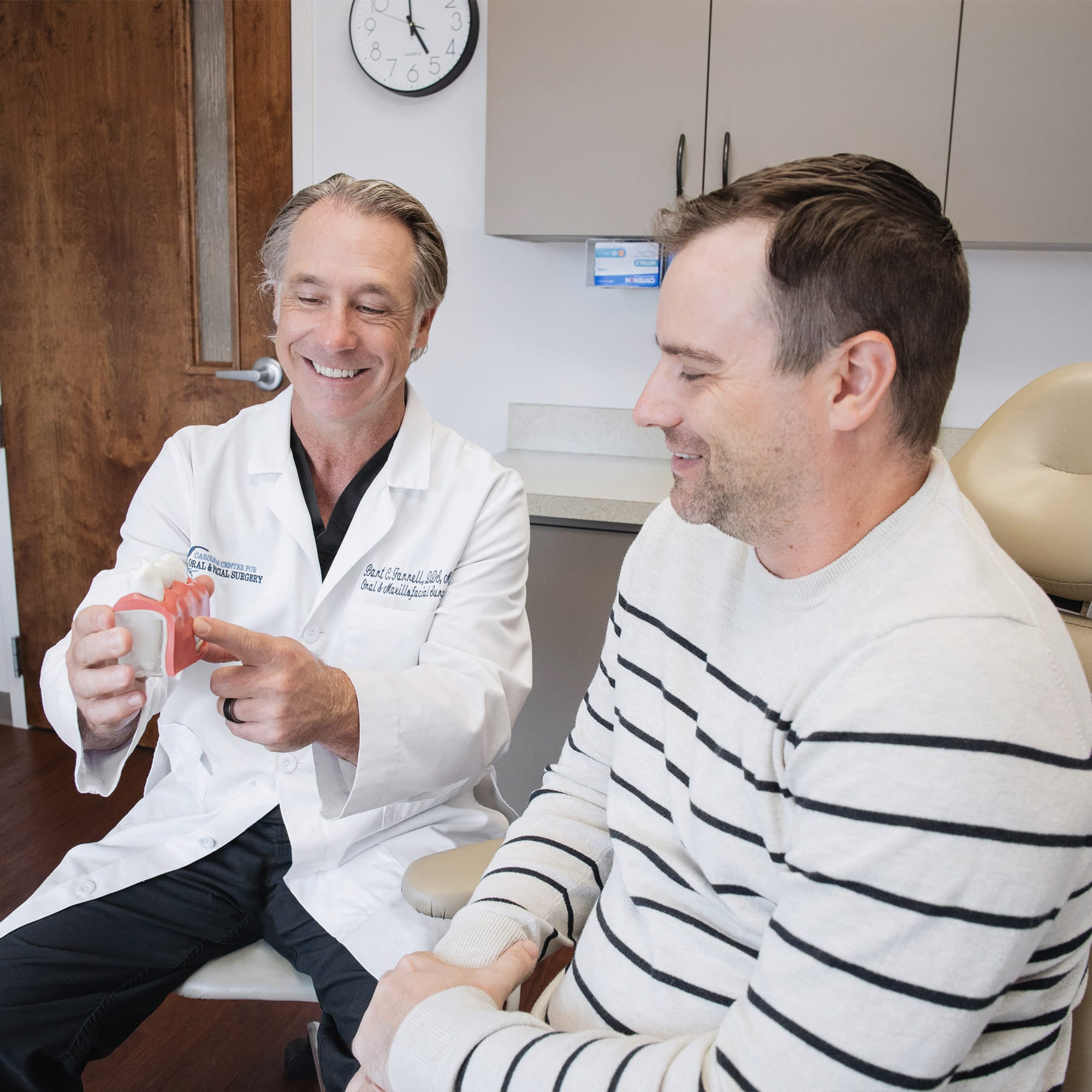 Two people in a medical office; one shows a dental model. Both are engaged and smiling. Wall clock and cabinets visible.
