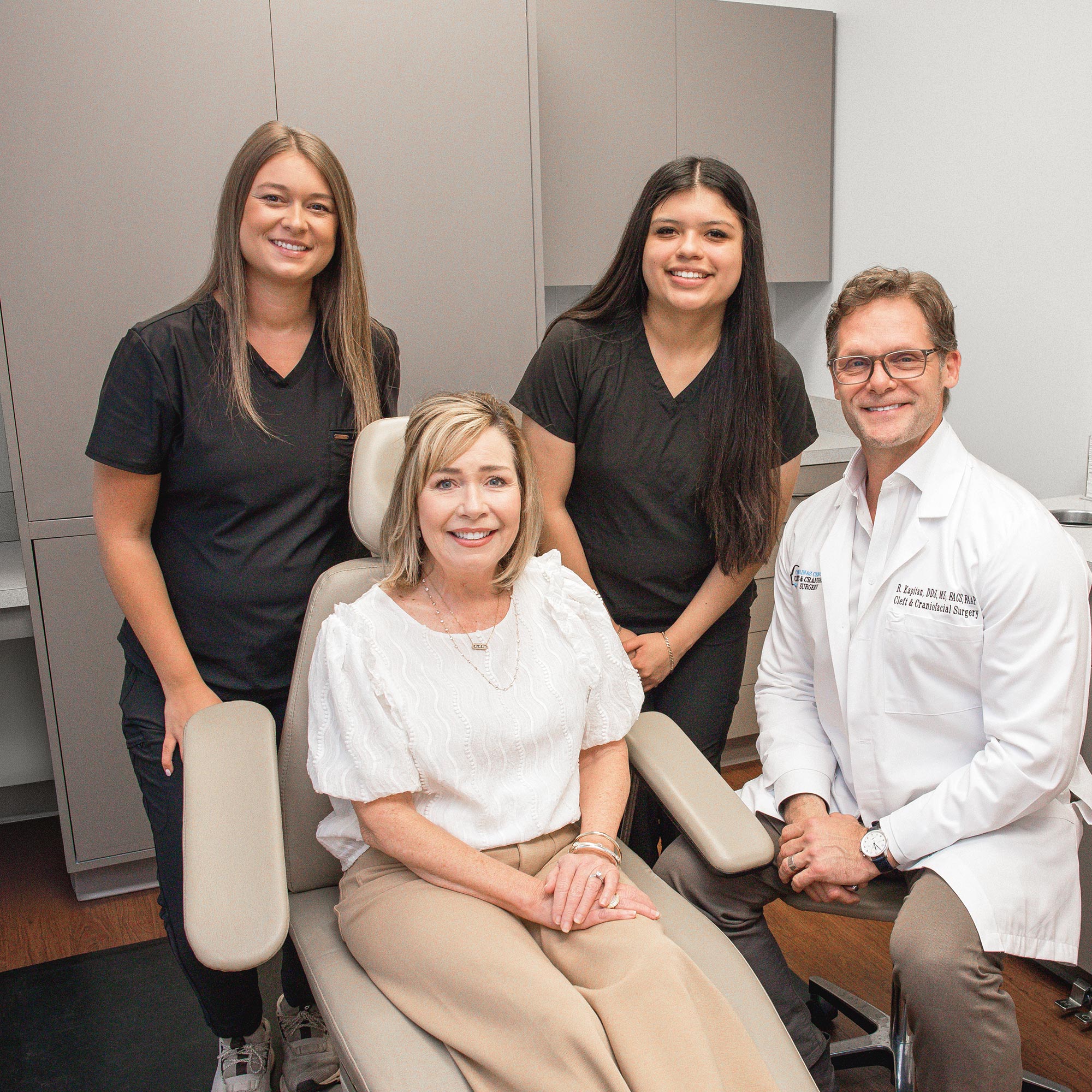 Four people are smiling in a medical office setting. A person is seated on a chair, surrounded by three standing individuals.