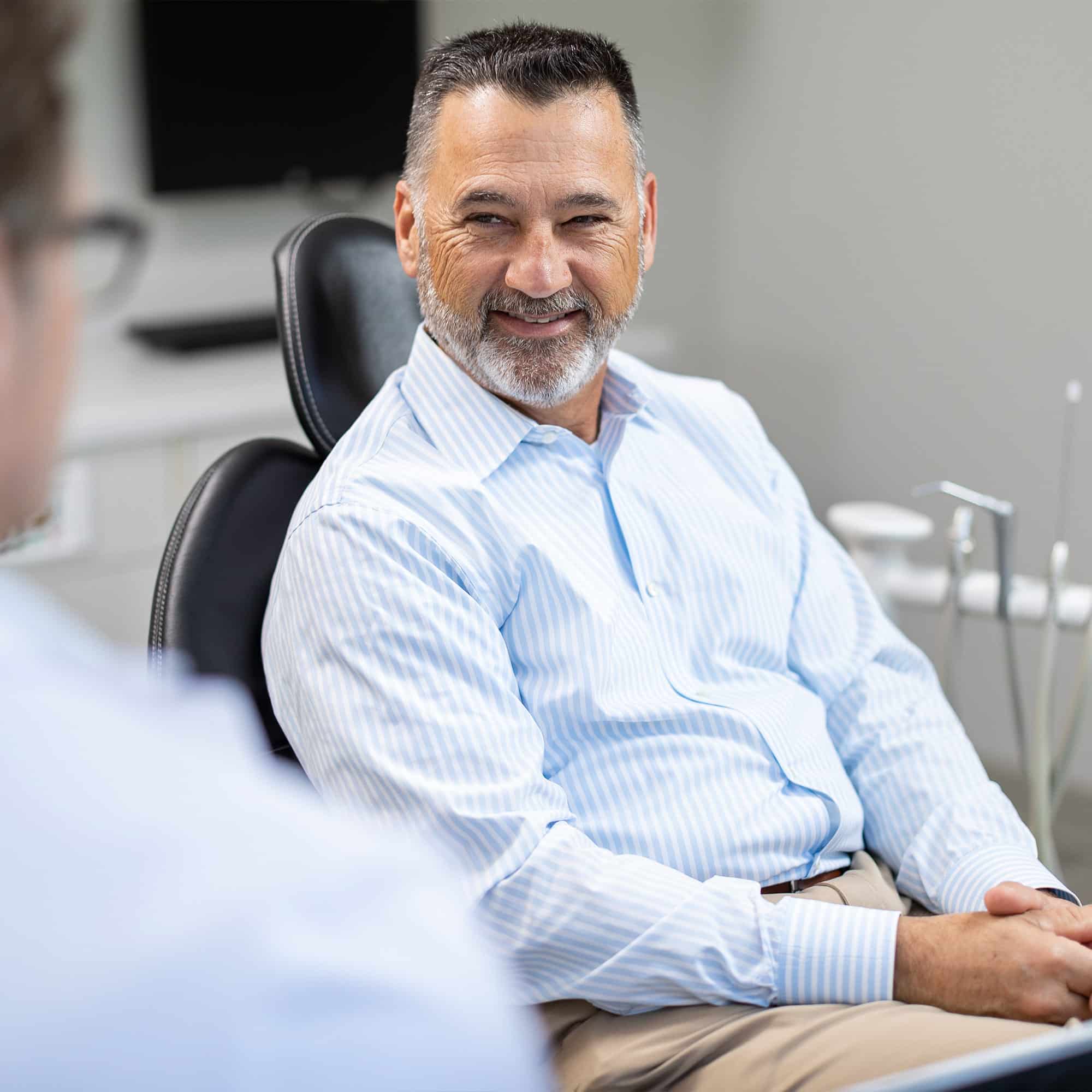 A smiling person sits in a dental office, wearing a striped shirt, engaging with another person. Dental equipment is visible in the background.