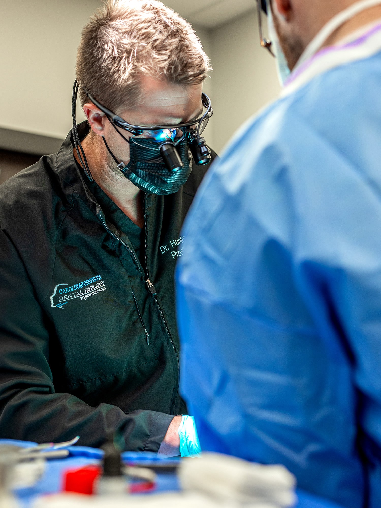 Two dental professionals in medical attire focus on a patient procedure, equipped with surgical eyewear and masks, in a clinical setting.