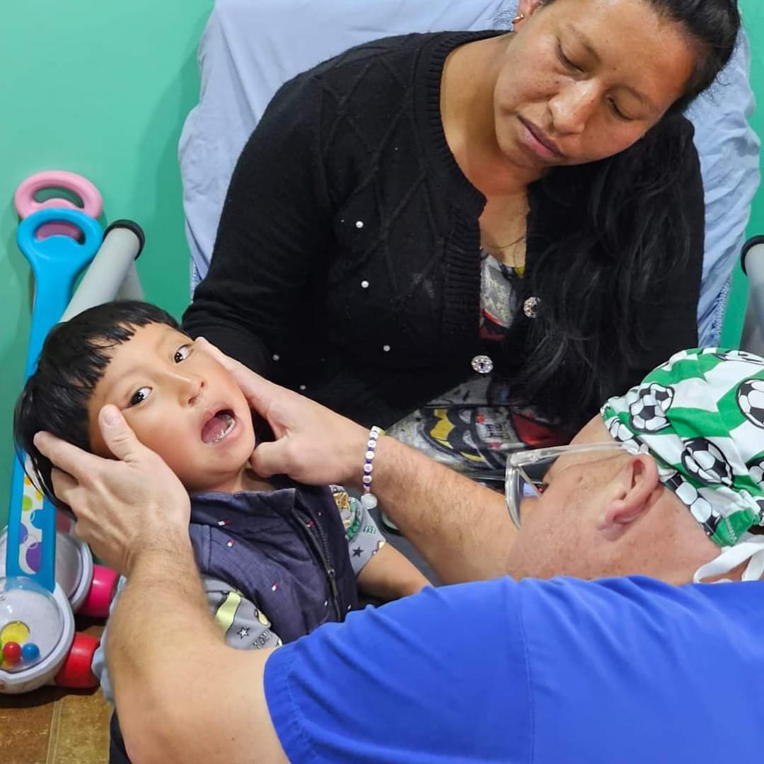 A person examines a child's mouth, while another person observes in a room with colorful toys in the background.