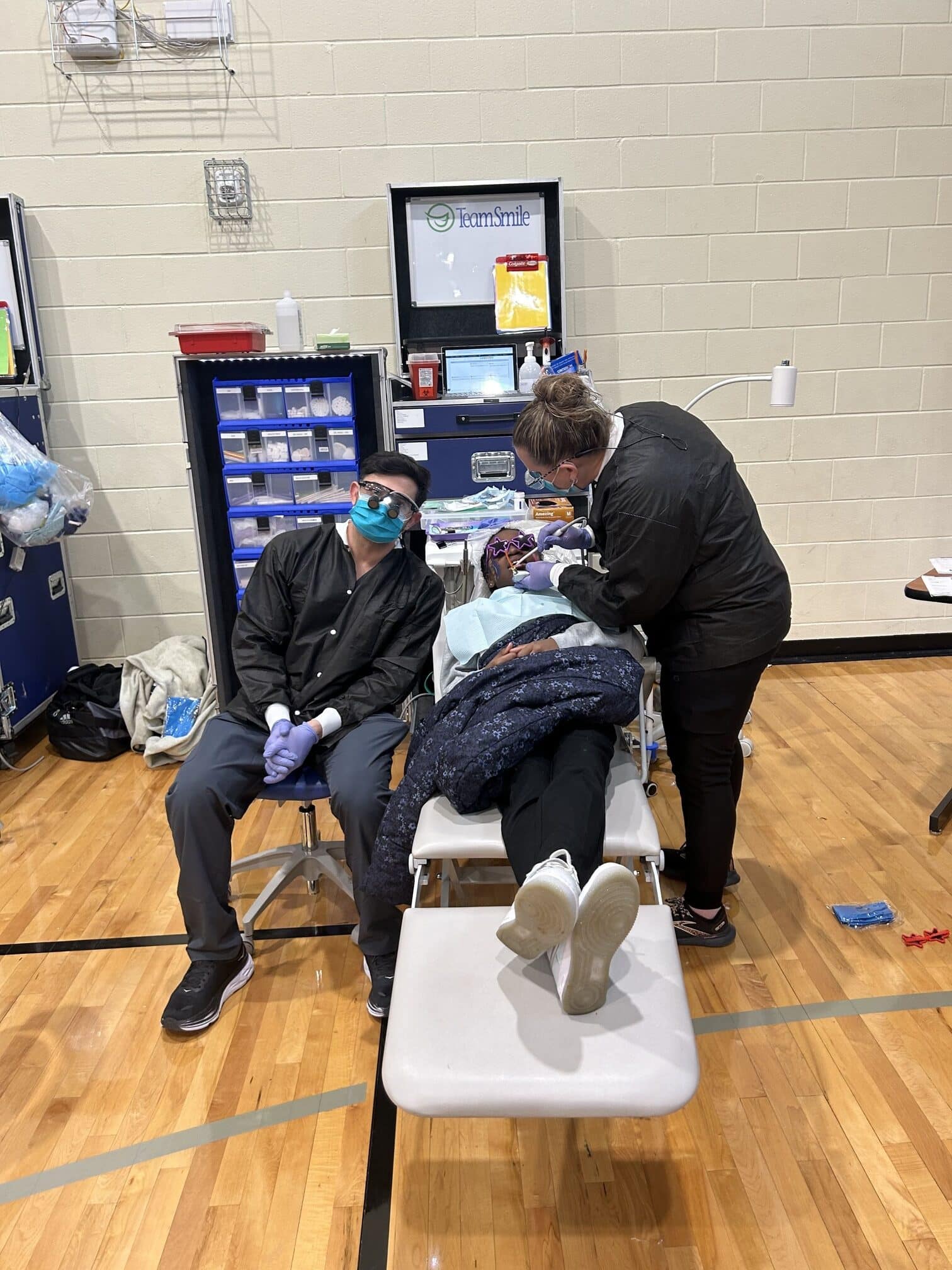 A person receives dental care in a gymnasium setting. Medical equipment is organized nearby. "Team Smile" is visible on a cabinet.