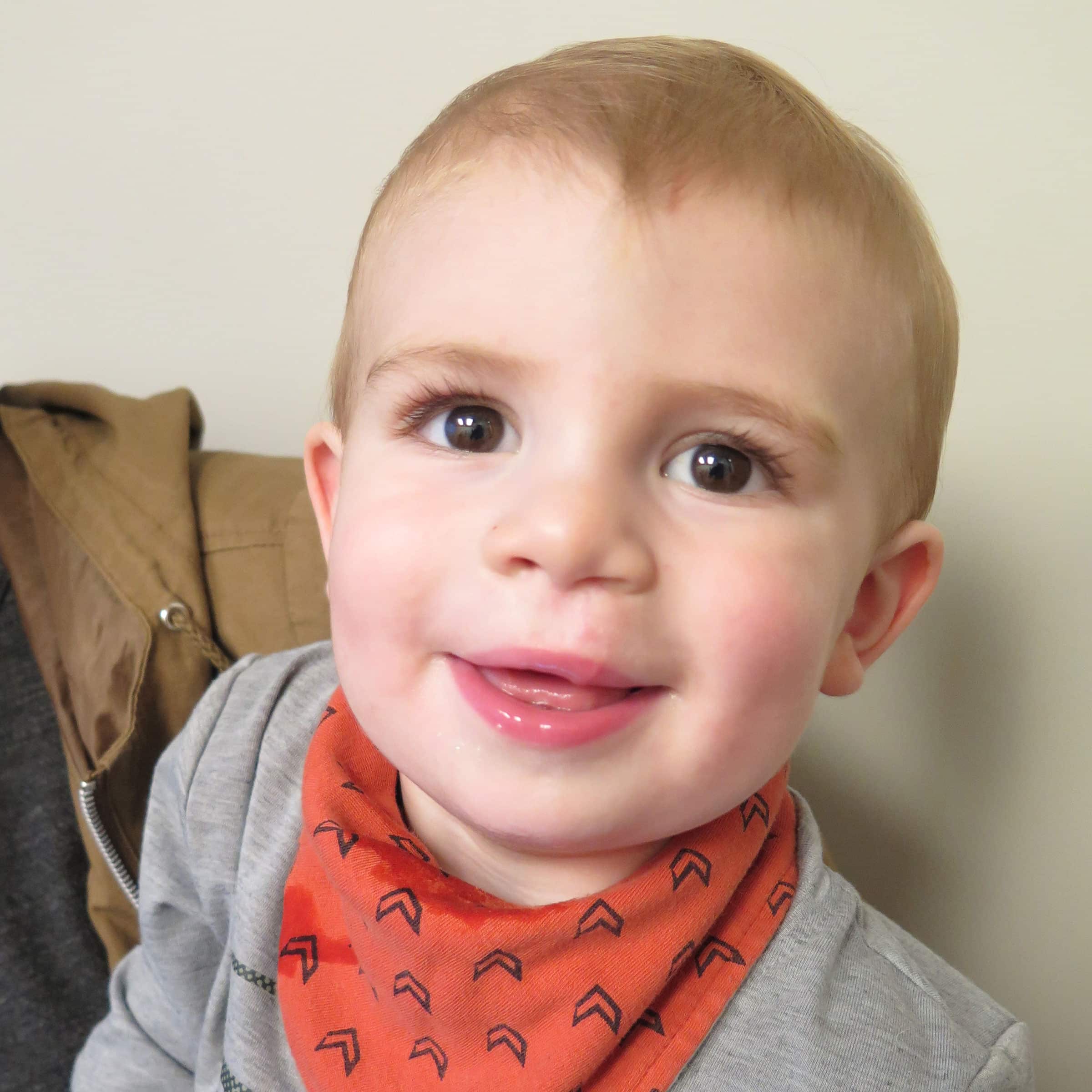 A young child with an orange patterned bib smiles warmly indoors, next to a person wearing a jacket. Neutral background setting.