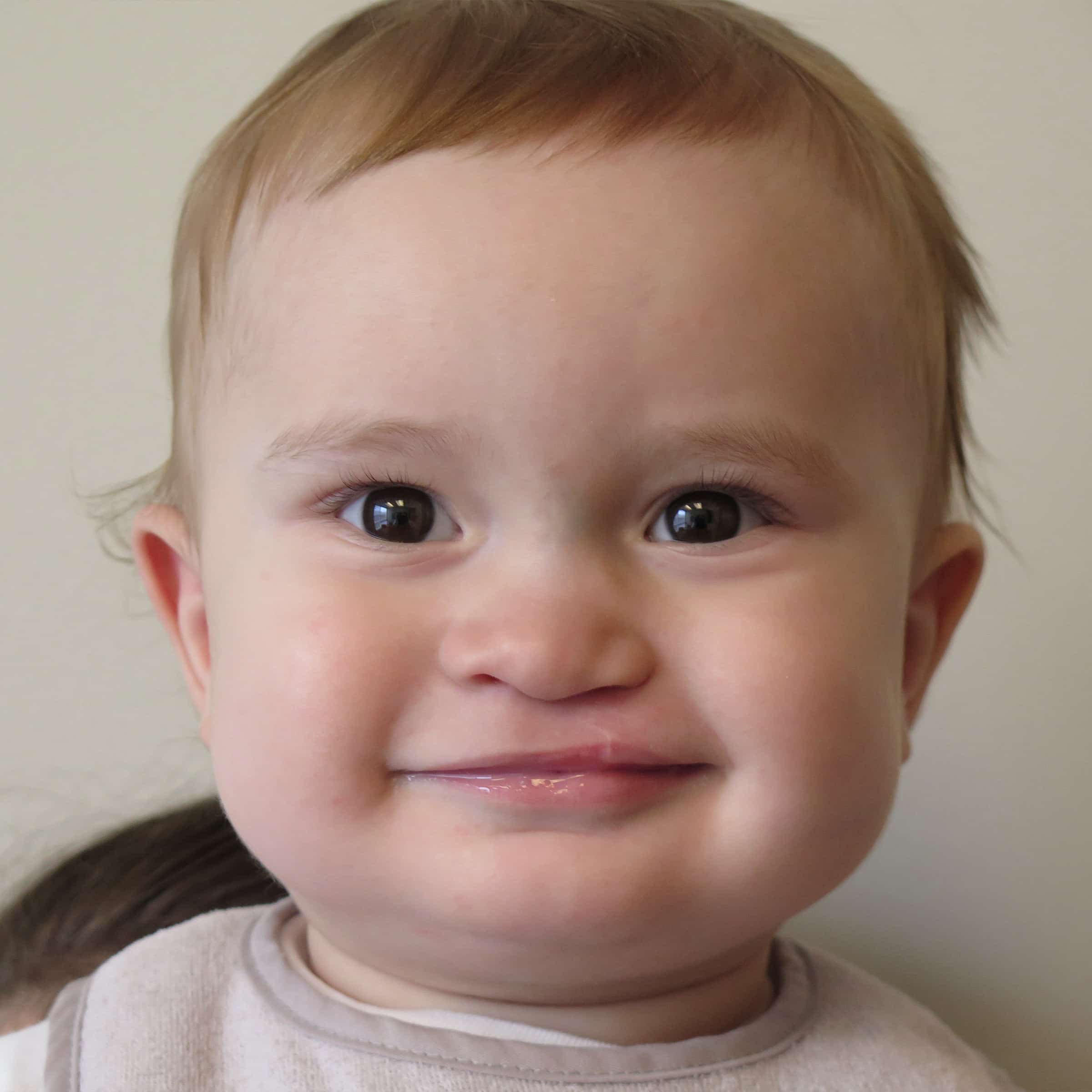 Close-up of a smiling baby with light hair against a plain background. The baby has bright eyes and a joyful expression.