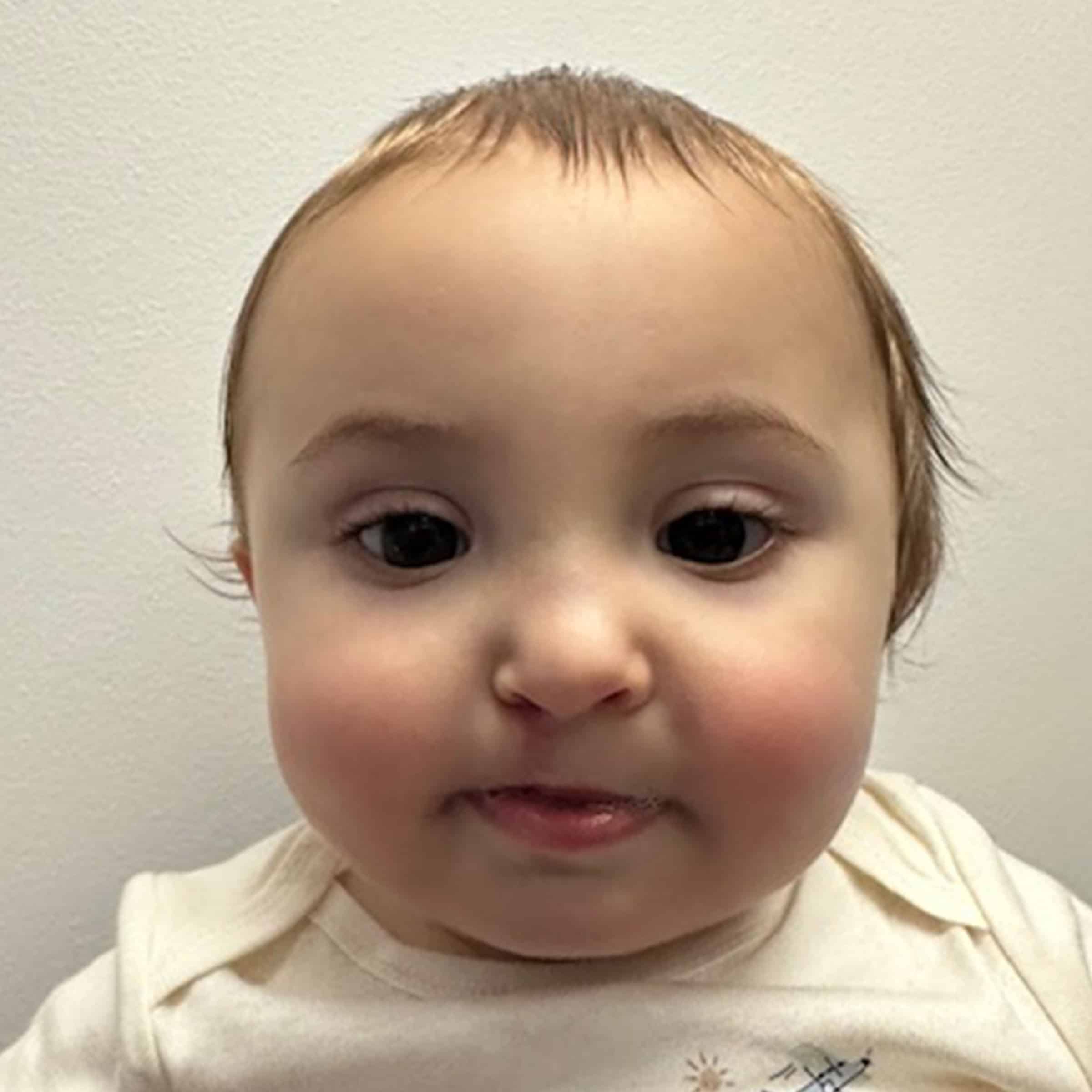 A baby with light hair and big eyes, wearing a cream-colored shirt, looks directly at the camera against a plain white background.