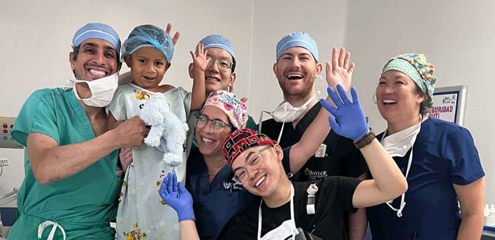 Group of smiling medical professionals and a child in scrubs, posing cheerfully in a clinical setting.