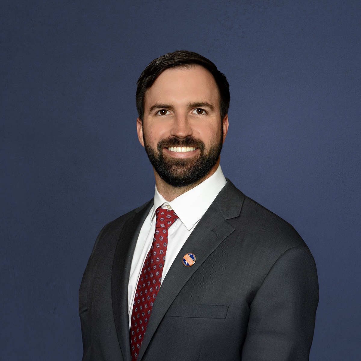 A person in a suit and red tie smiles against a plain blue background, wearing a pin on the lapel.