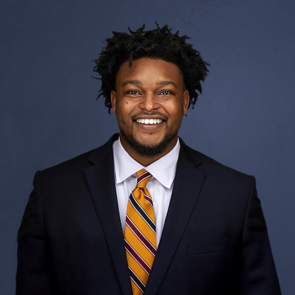 A person in a suit with a striped tie is smiling against a plain dark blue background.