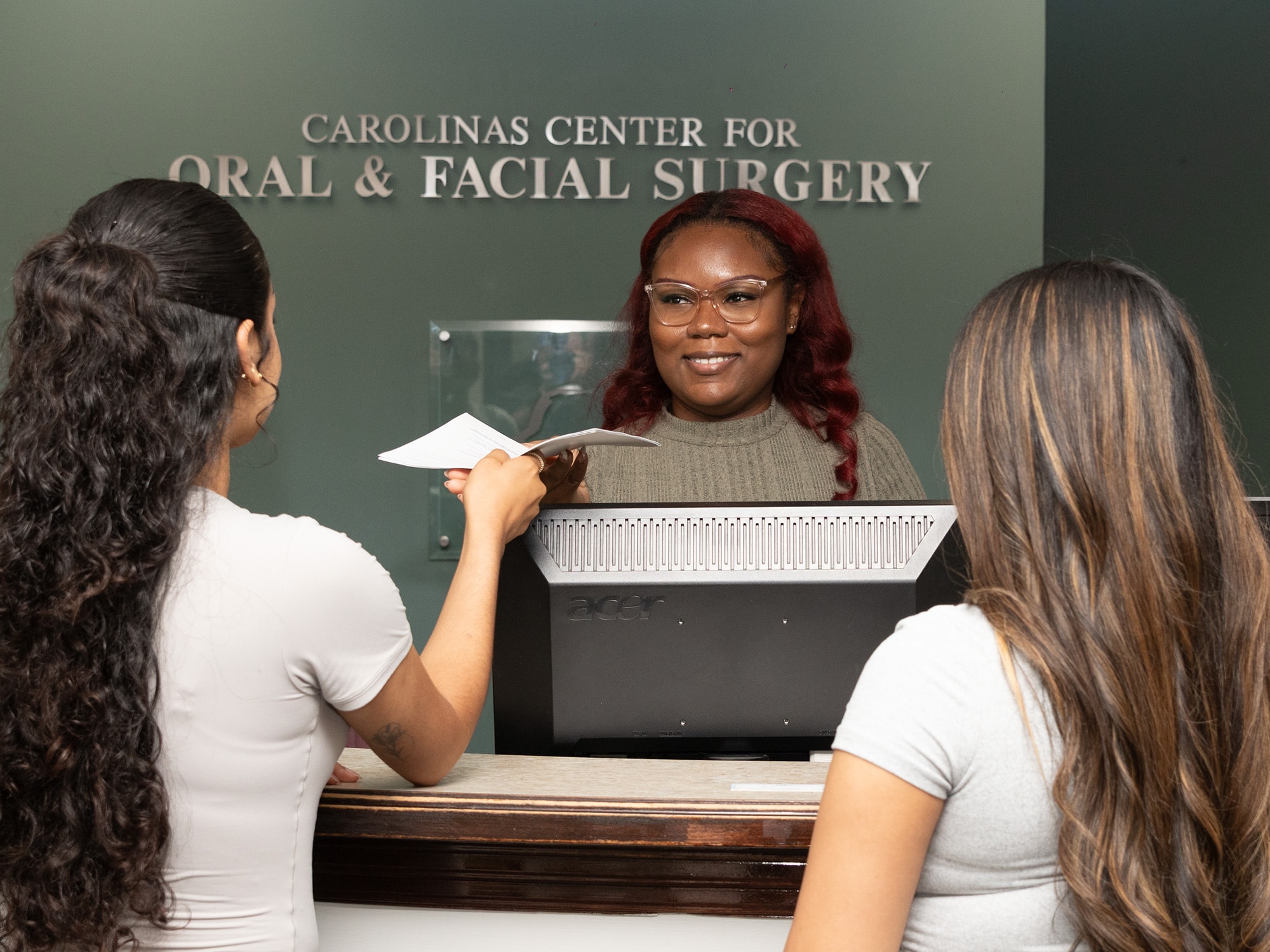 Three people interact at the reception of Carolinas Center for Oral & Facial Surgery, with one person behind a computer receiving documents.