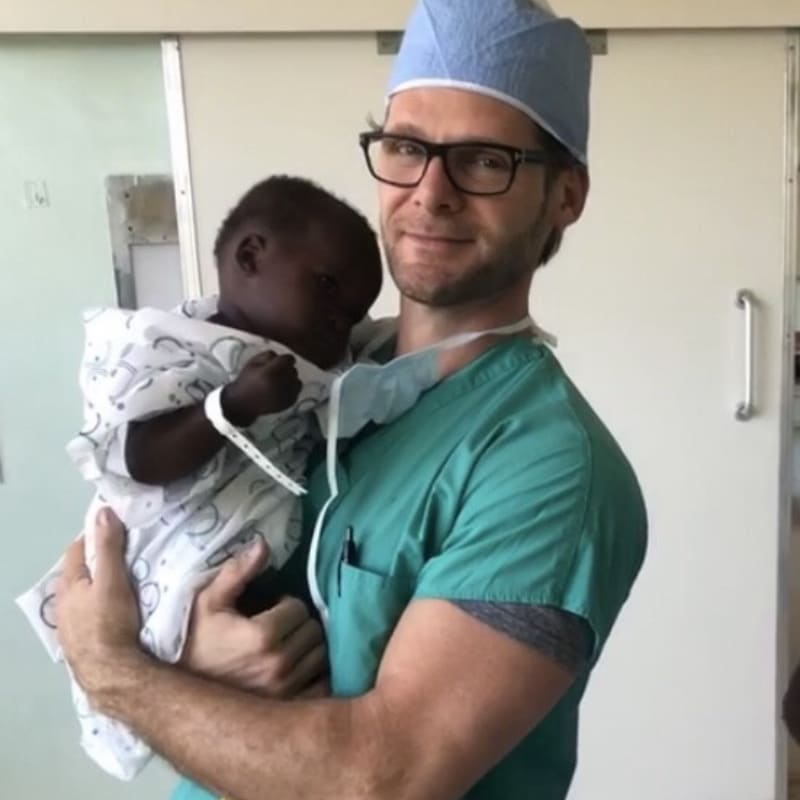 A person in medical scrubs holds a baby in a hospital room, smiling gently at the camera.