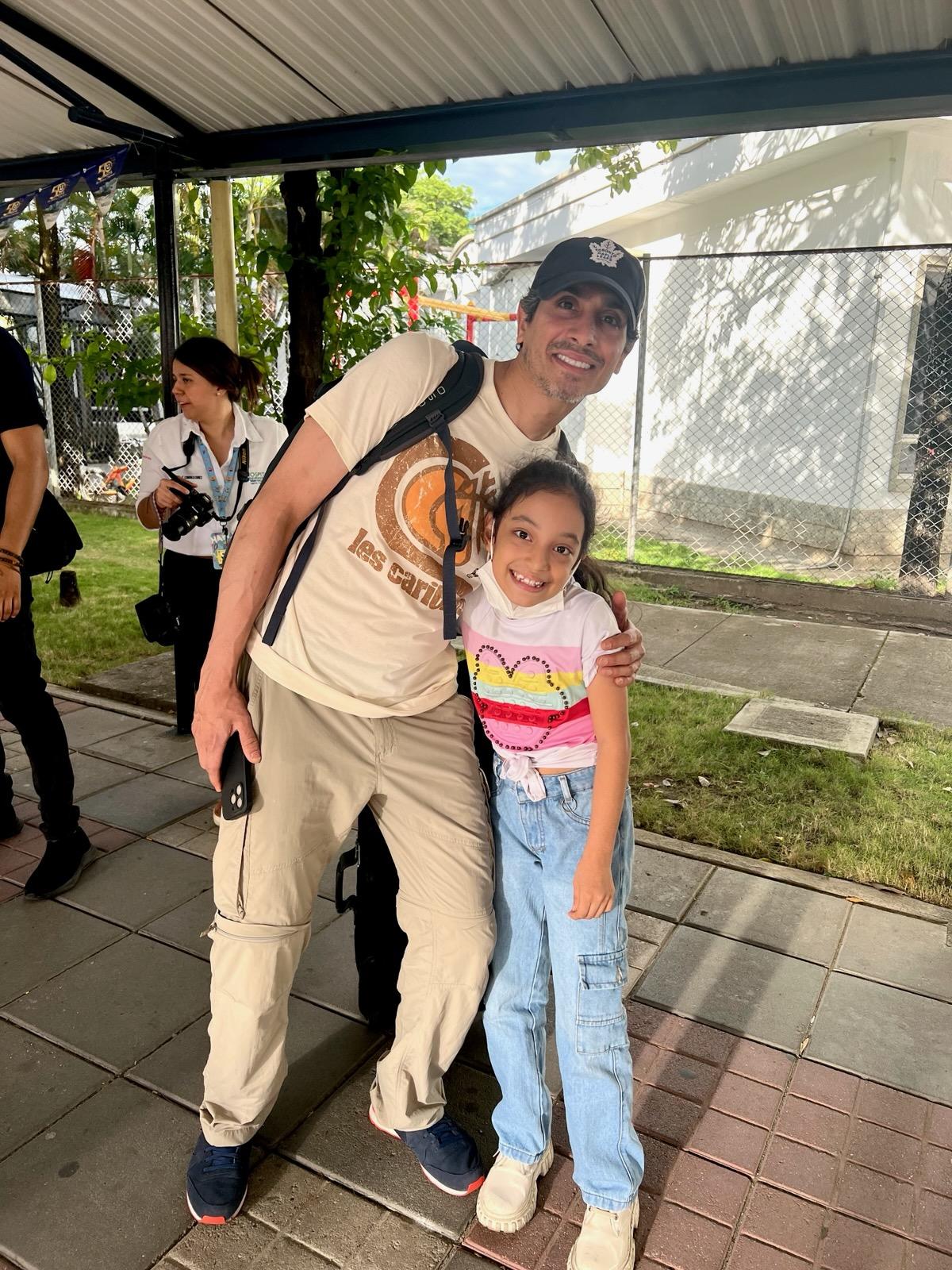 A person and child smile under a metal canopy. Another person in white stands nearby. There's greenery and a chain-link fence in the background.