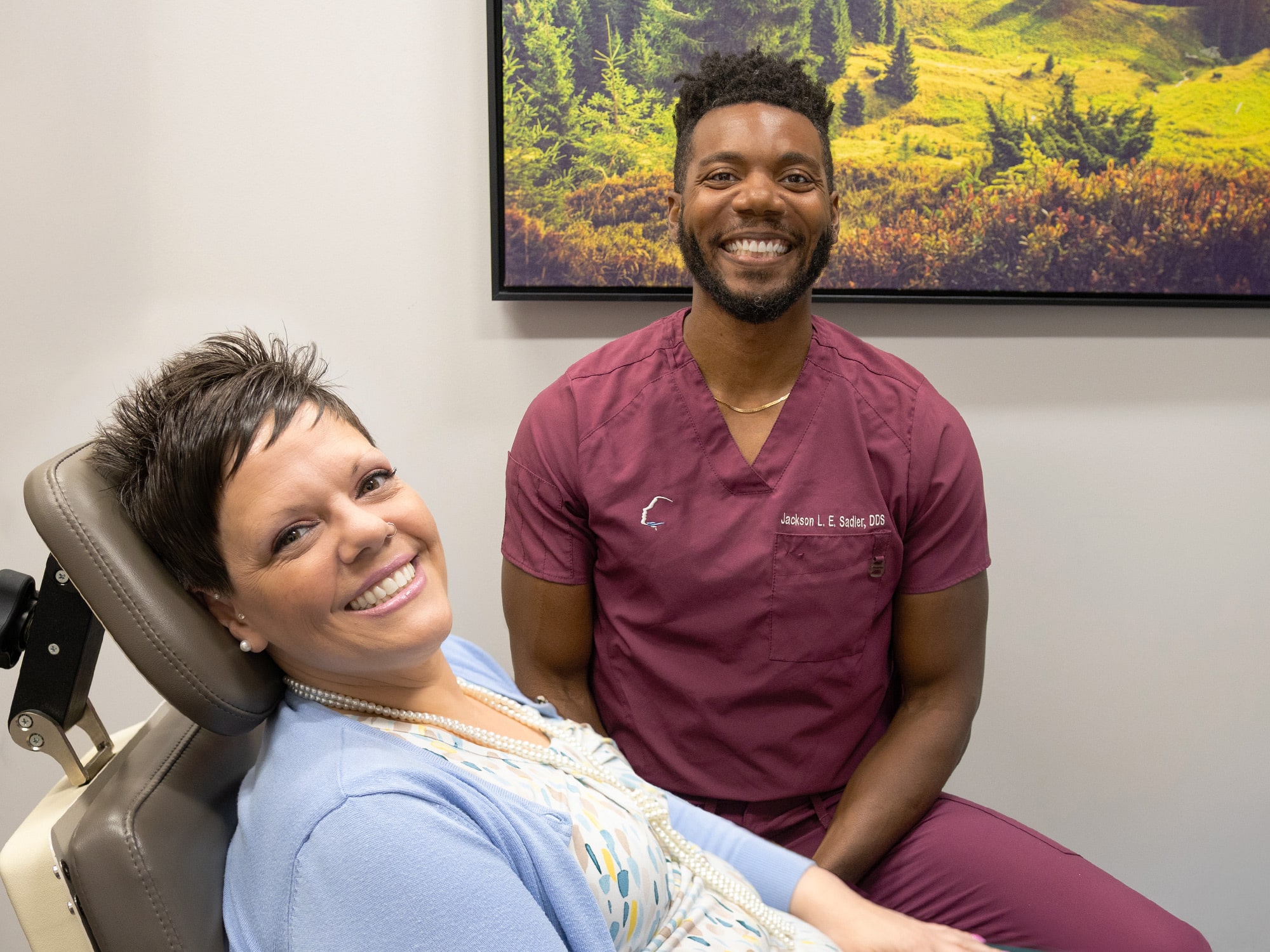 Two smiling people in a dental office setting, person in scrubs, patient reclining in chair, vibrant landscape artwork on wall.