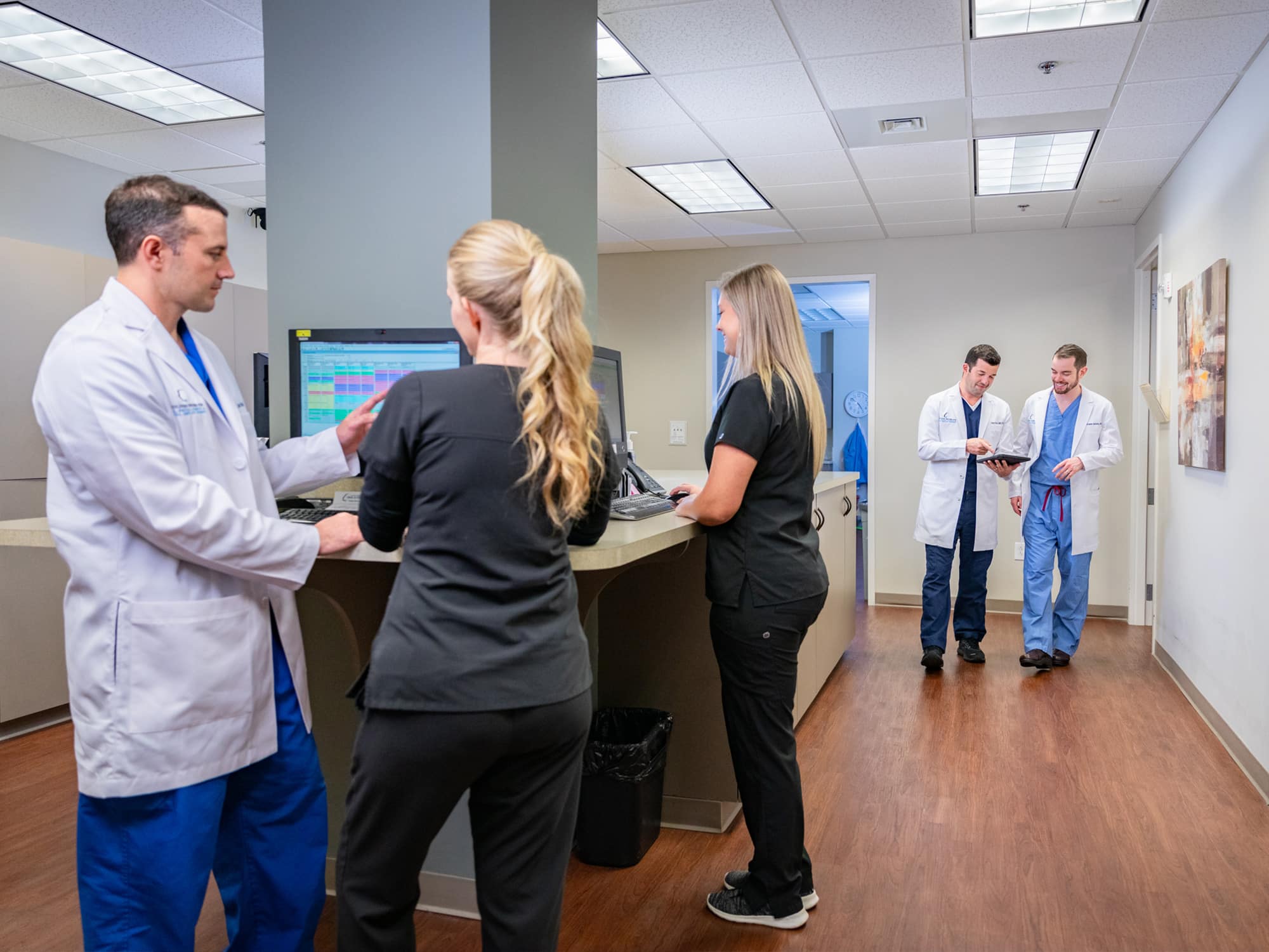 Four medical professionals in scrubs and coats converse in a brightly lit hospital corridor with computers and a modern design.