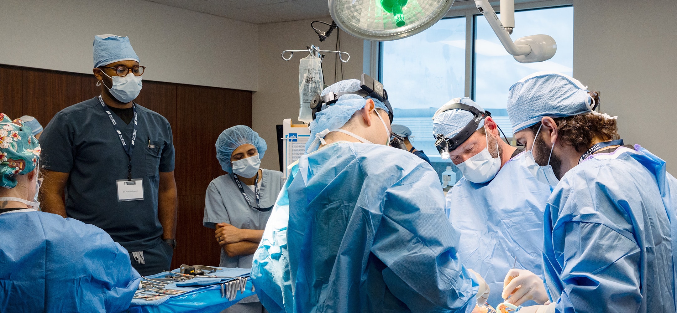 Medical team in surgical attire performs operation in hospital operating room under bright surgical light, with various surgical instruments on table.